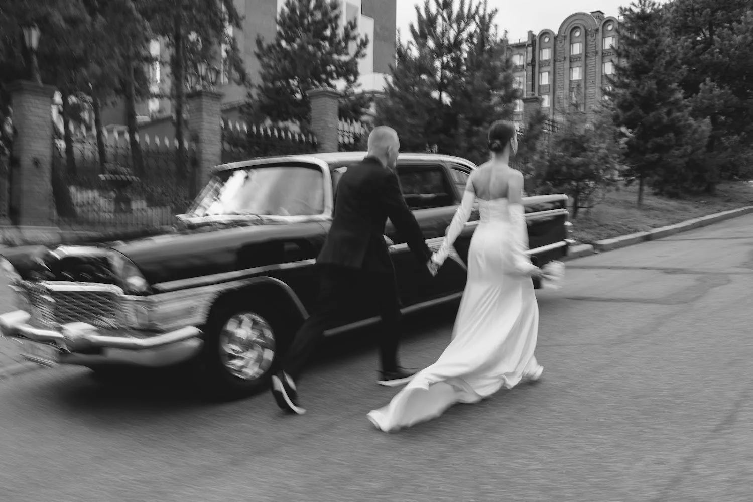 A black-and-white photo of a couple holding hands and walking away from a vintage black car on a city street, with trees and buildings in the background.