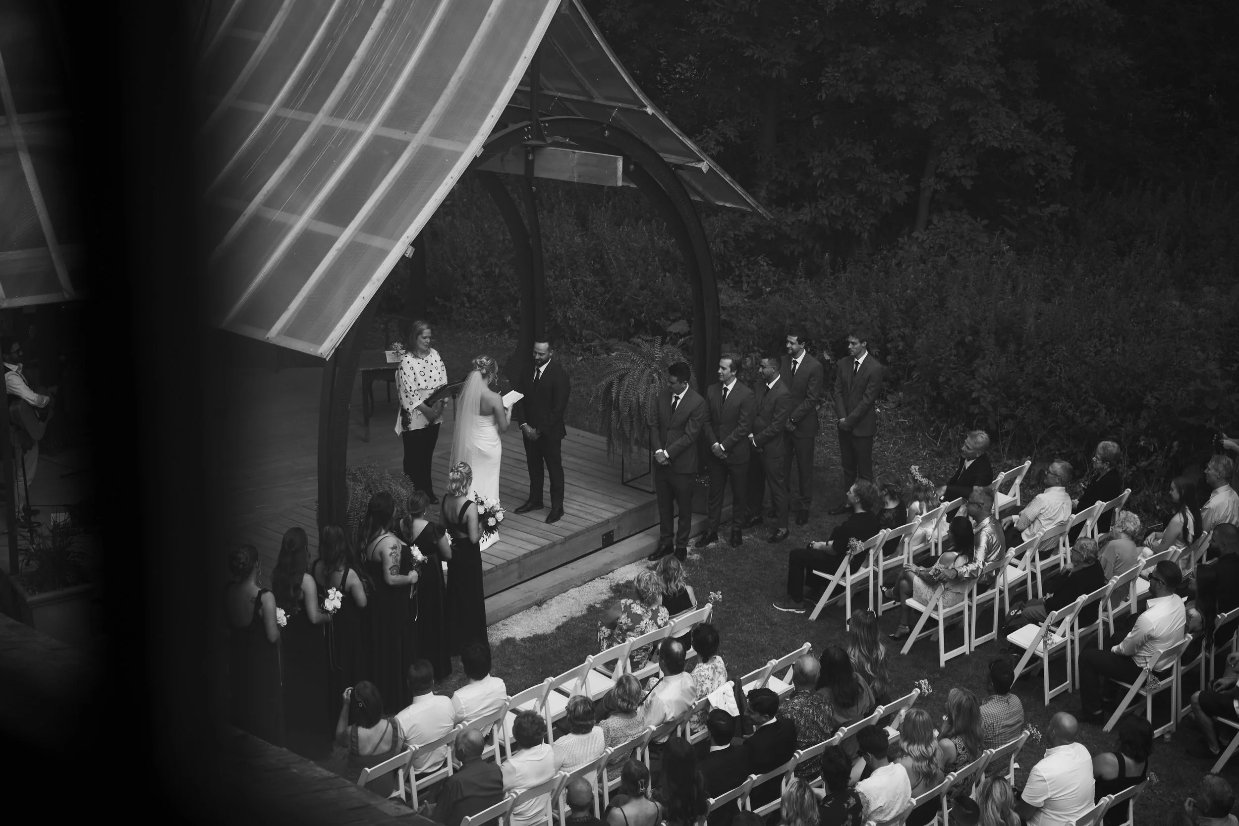 A black and white photo of a wedding ceremony outdoors with guests seated and a couple exchanging vows on a small wooden platform under a canopy.