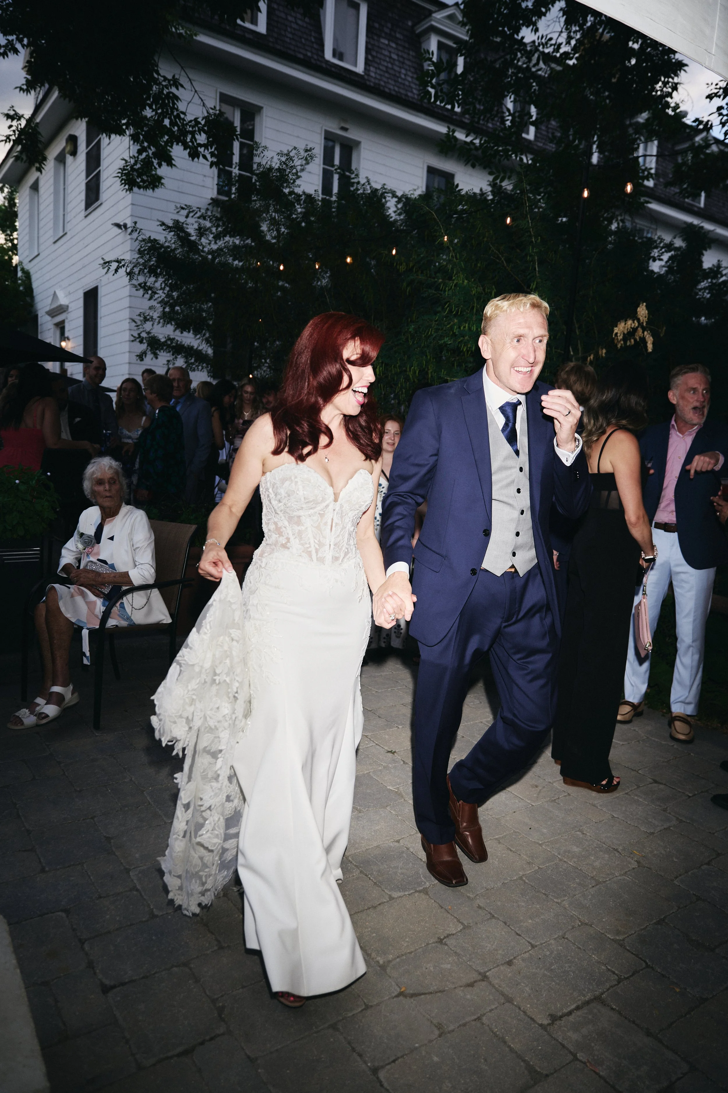 A bride and groom walking hand in hand at their wedding reception, smiling, with guests in the background outdoors during evening.