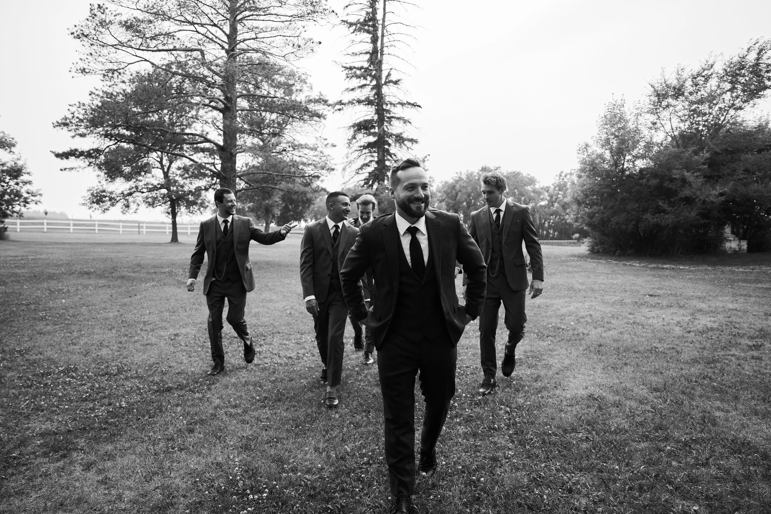 A group of six men dressed in suits walking outdoors on a grassy field, some smiling and chatting, with trees and a white fence in the background.