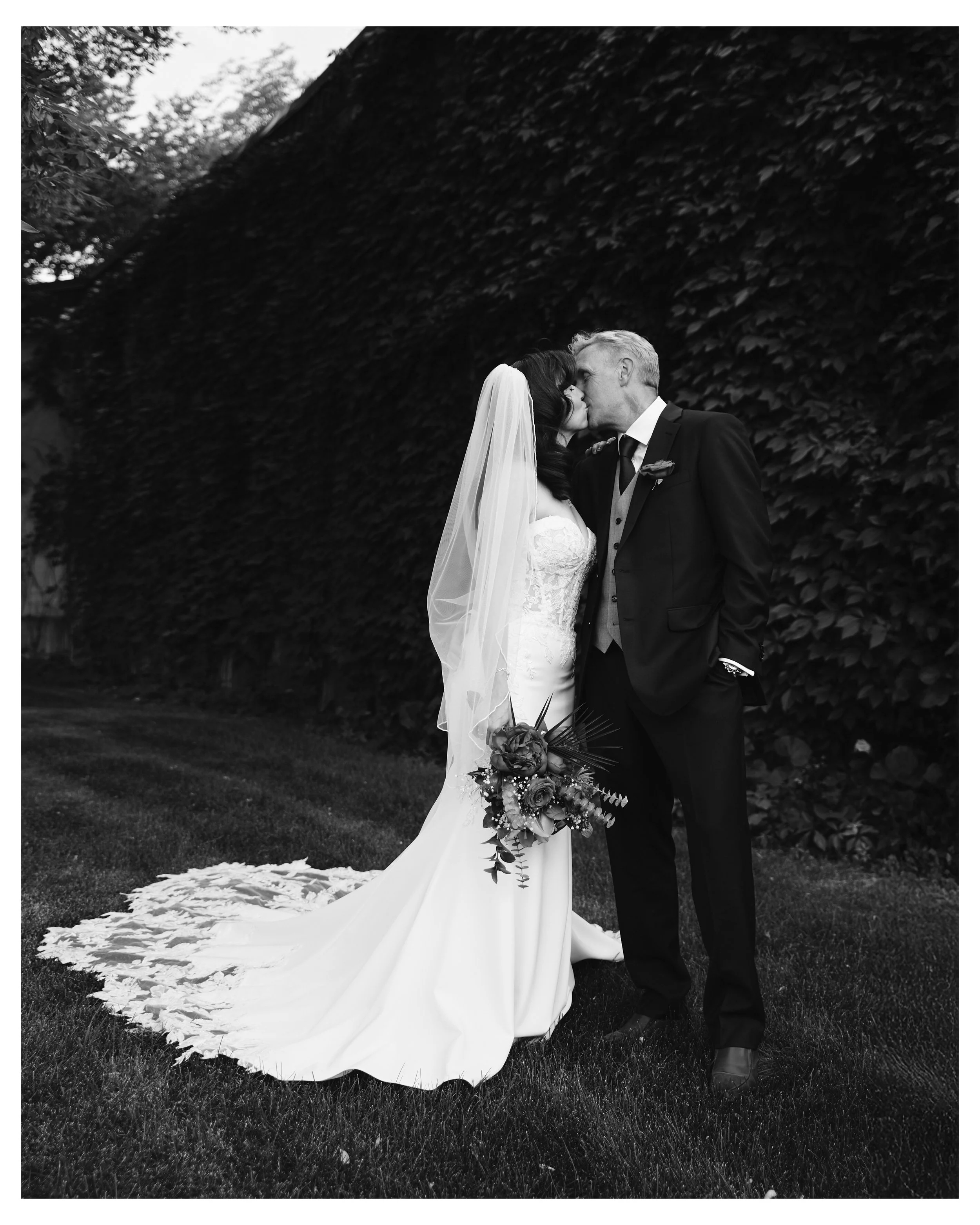 Black and white photo of a bride and groom sharing a kiss outdoors. The bride wears a lace wedding gown and veil, holding a flower bouquet, while the groom is dressed in a suit with a boutonniere. They stand on grass with a large leafy hedge behind t