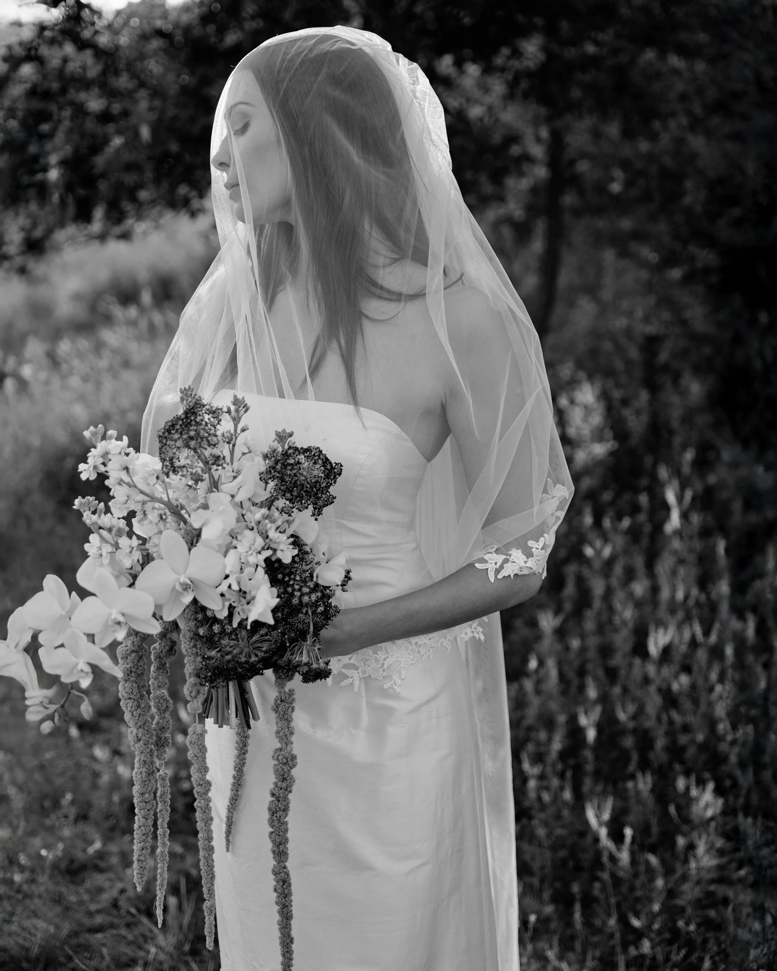 A woman in a wedding dress with lace details on the sleeves, wearing a veil, is holding a bouquet of flowers and standing outdoors near trees.