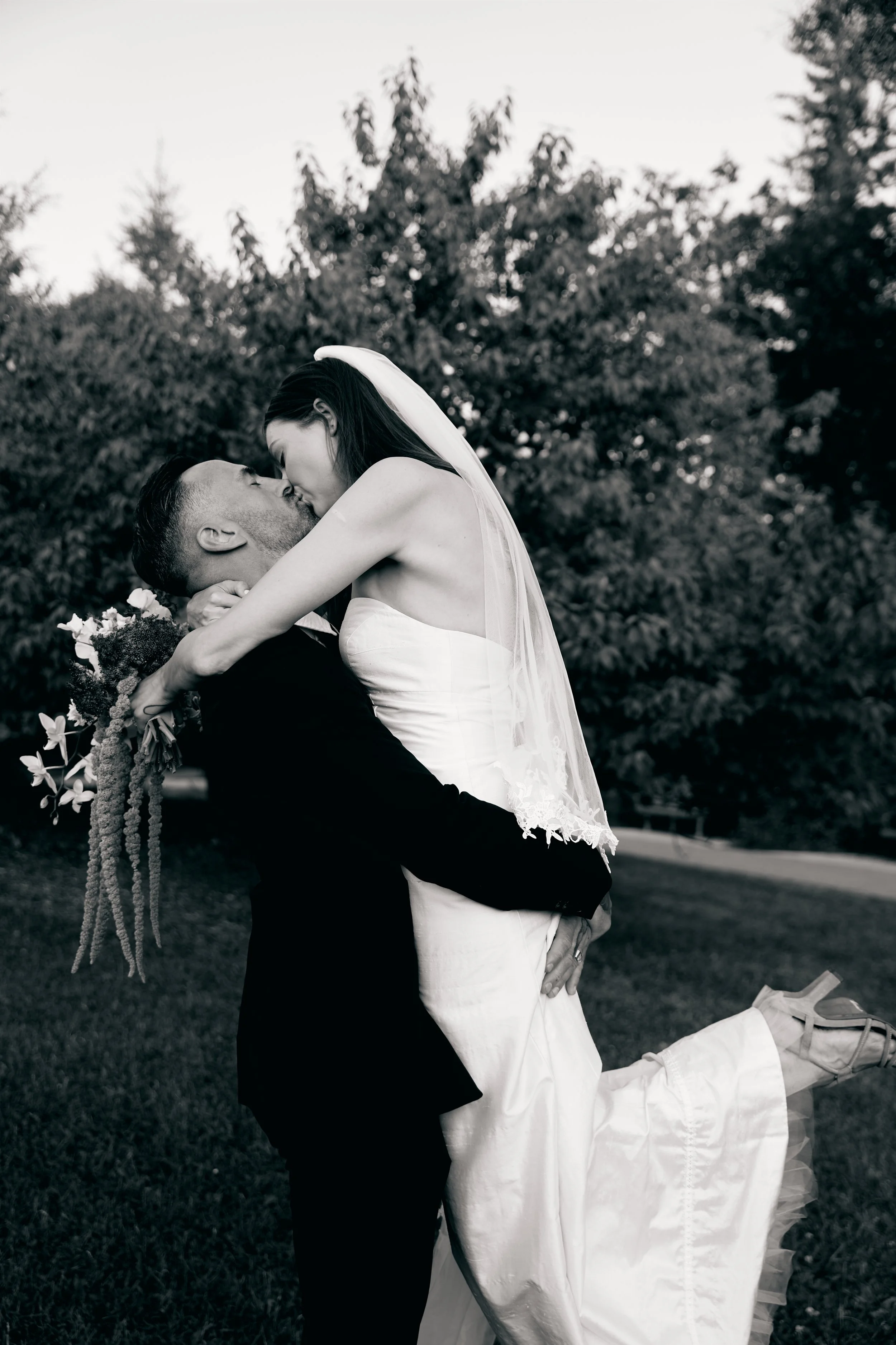 A black and white photograph of a bride in a white wedding dress and veil lifting her leg as she kisses a groom who is holding her up outdoors with trees in the background.