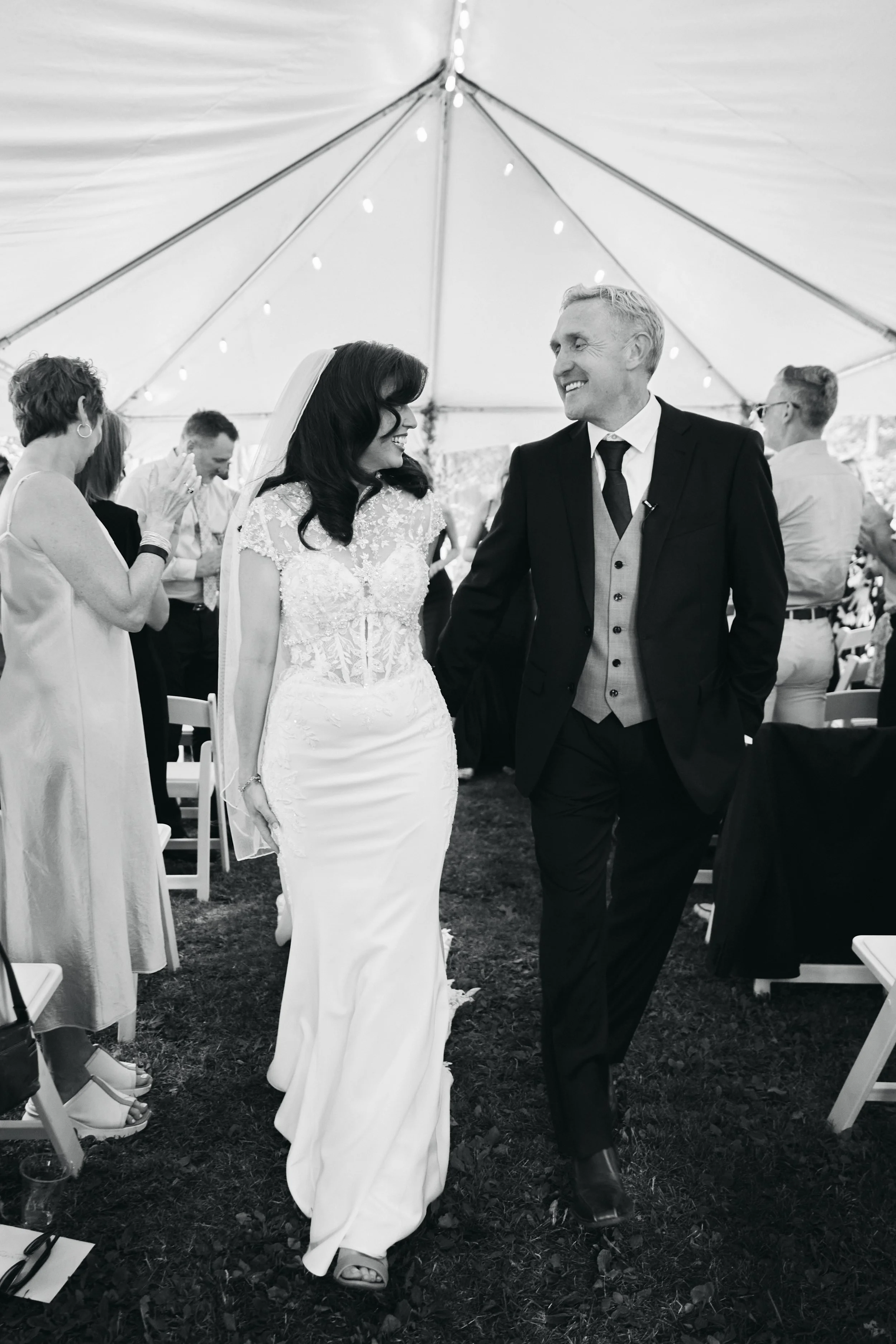 A bride and groom holding hands and smiling at each other during their wedding reception under a tent, surrounded by guests.