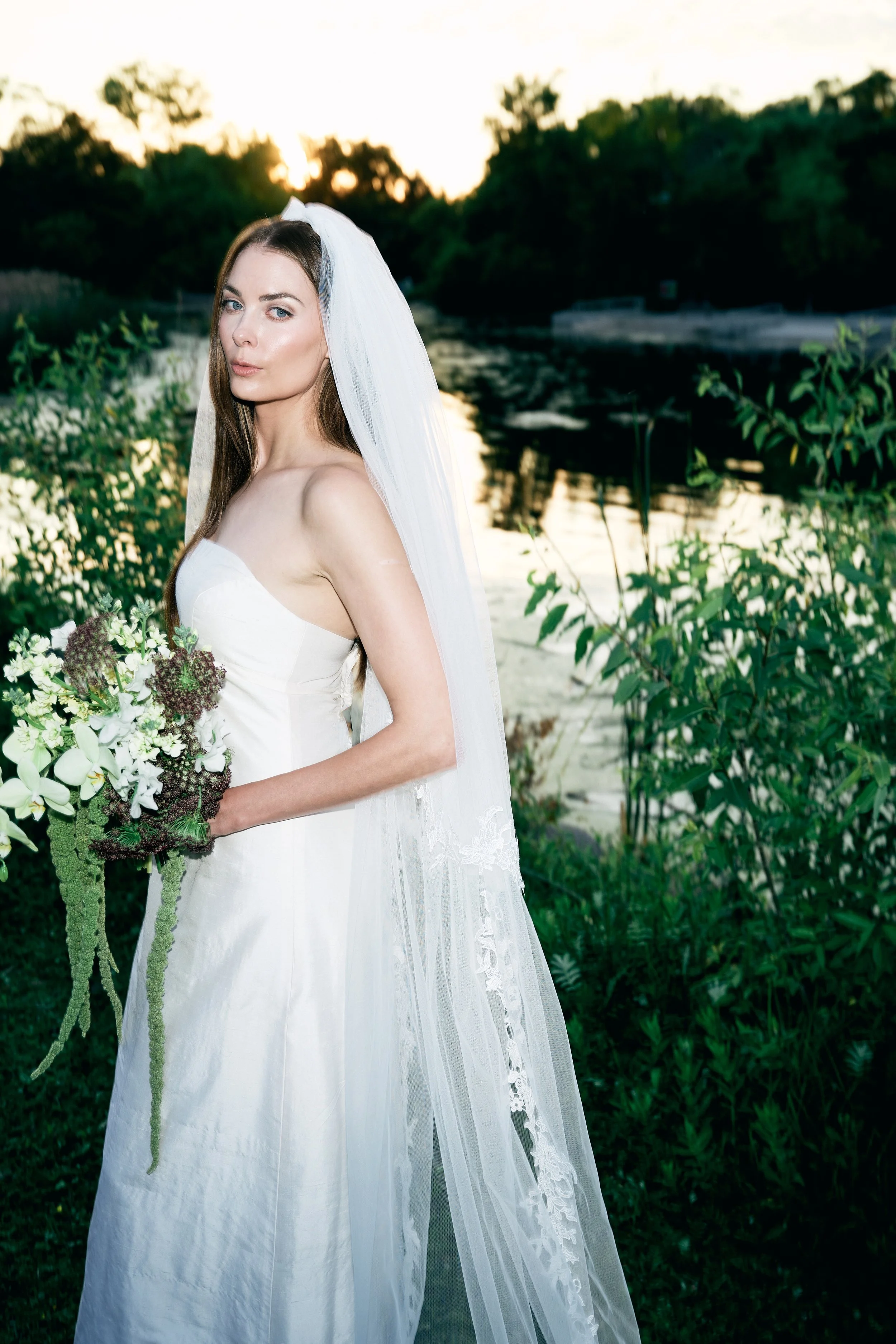 A bride in a white strapless wedding dress and veil holding a bouquet of flowers, standing outdoors near a body of water at sunset.