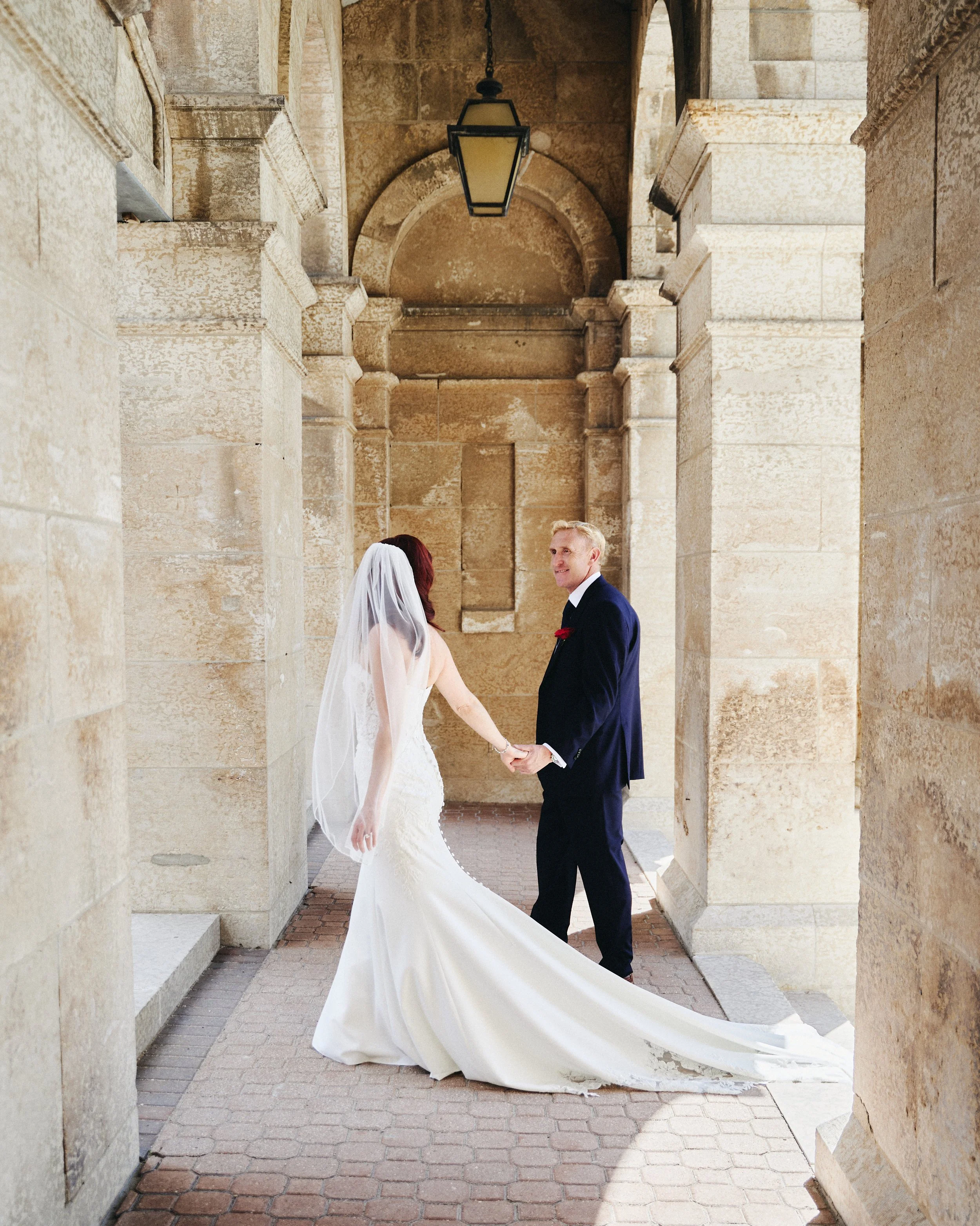 A bride and groom holding hands and facing each other in a historic stone archway.