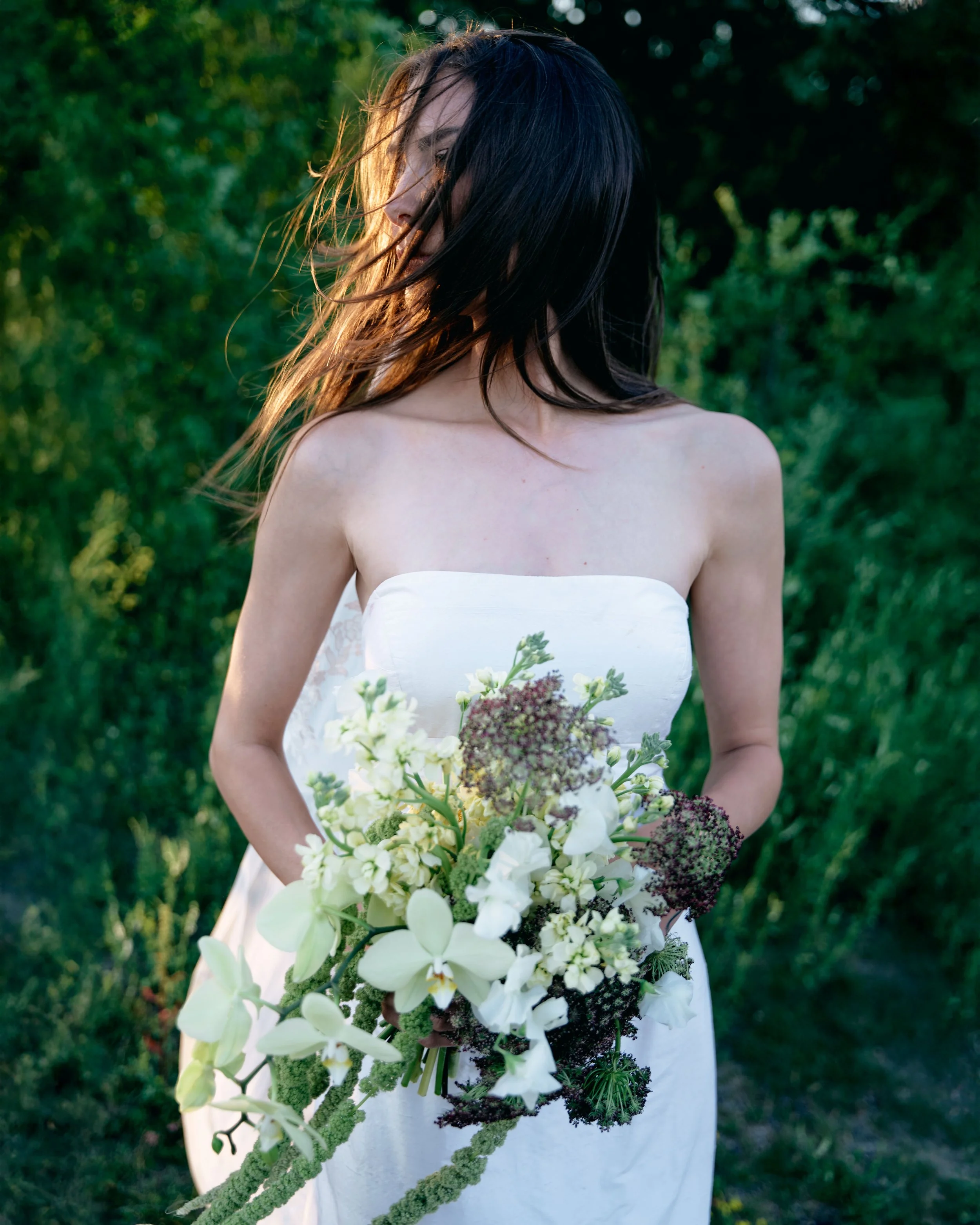 A woman in a white strapless dress holding a large bouquet of white and green flowers outdoors with greenery in the background.