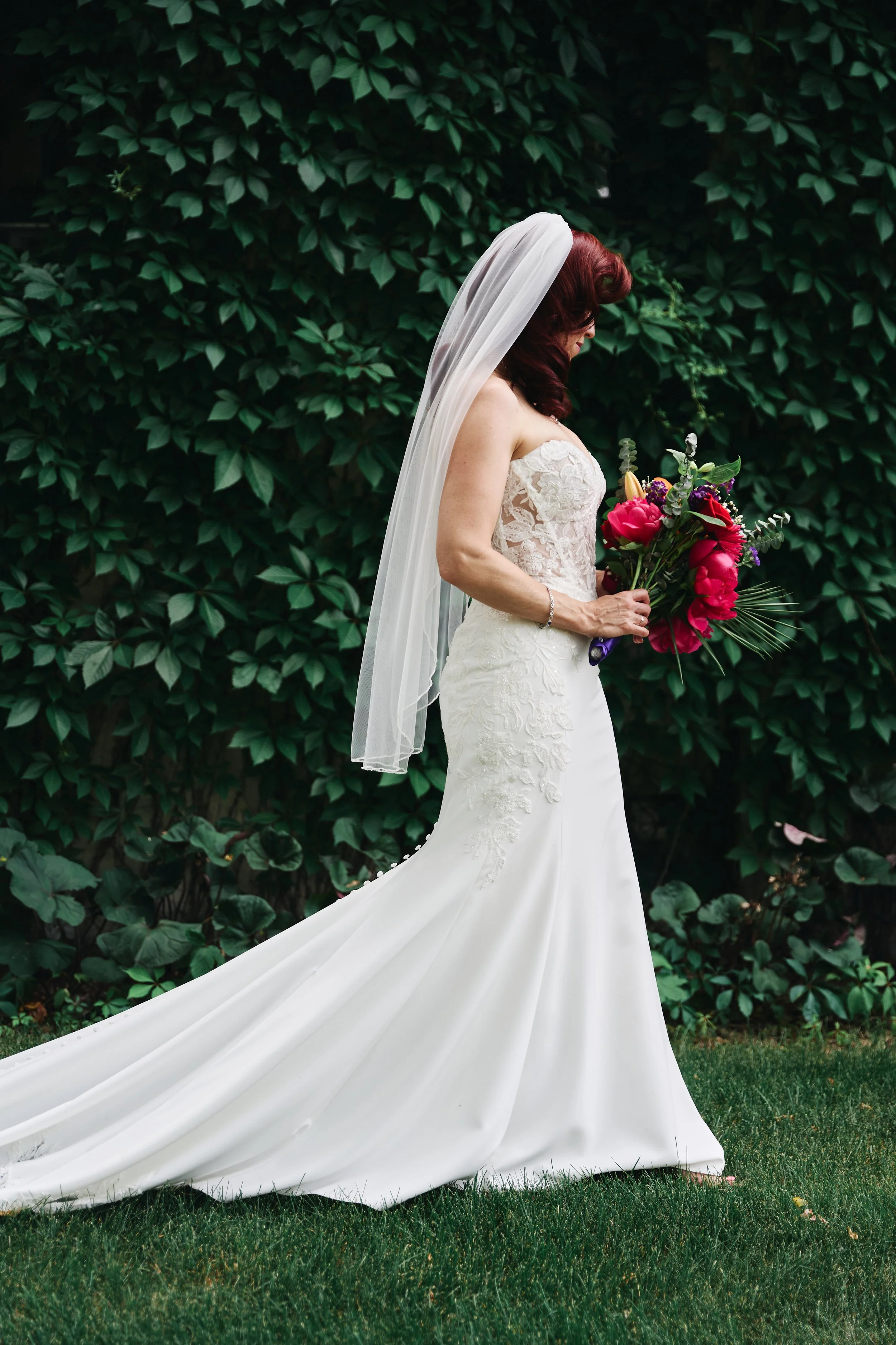 A bride in a white lace wedding gown holding a bouquet of pink, purple, and green flowers, standing outdoors on grass with a leafy green background.