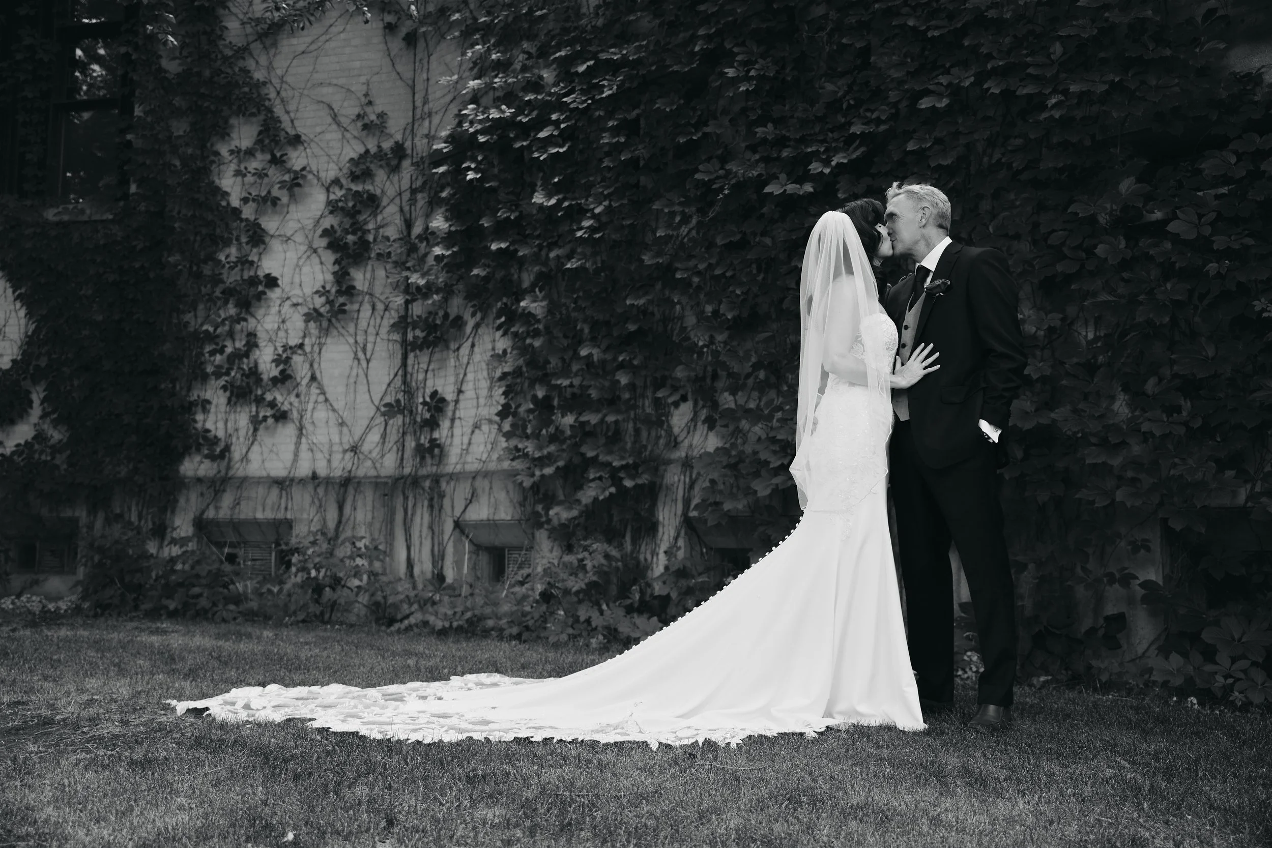 Black and white photo of a bride and groom sharing a kiss outdoors, with the bride wearing a wedding gown and veil, and the groom in a dark suit, standing on grass in front of a wall covered with ivy.
