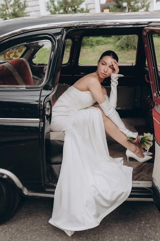 A woman in a white wedding dress sitting inside a vintage black car, holding a bouquet of flowers, with a pensive expression and her hand on her forehead.
