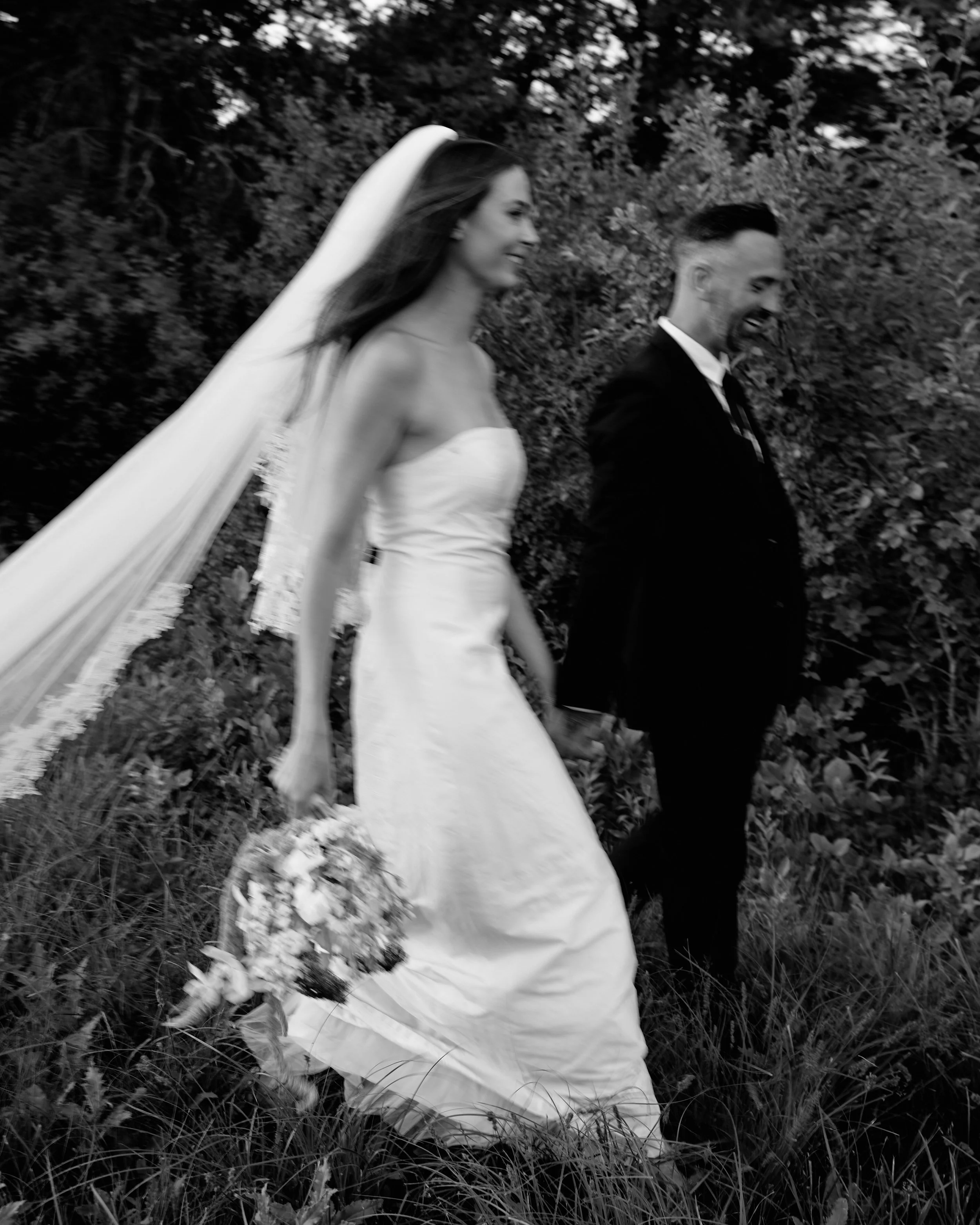 A bride and groom walking outdoors in a black and white photo, bride in a strapless wedding dress with a long veil holding a bouquet, smiling, and groom in a dark suit also smiling.