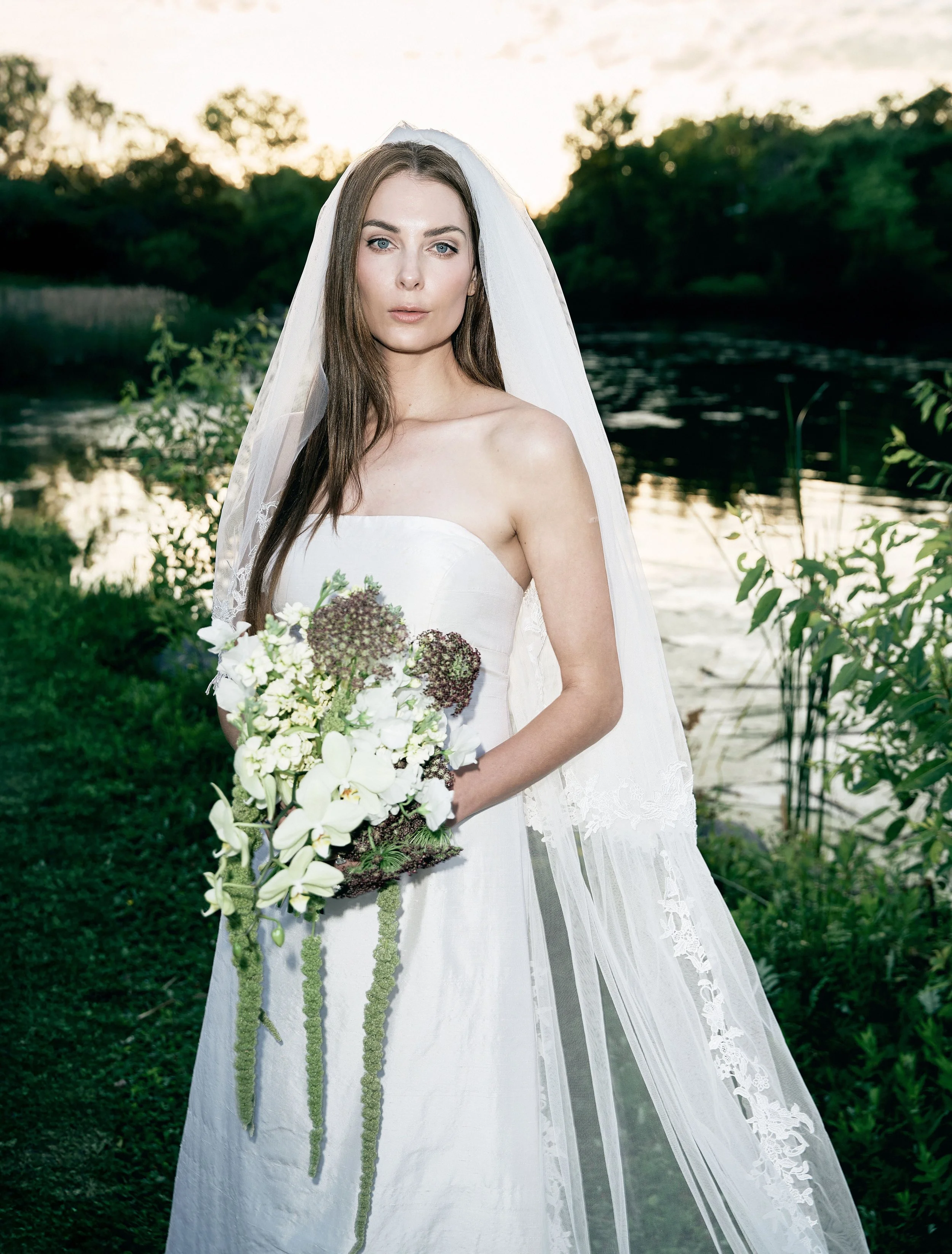 Bride in a white wedding dress holding a bouquet of white and green flowers standing outdoors near a body of water at sunset.