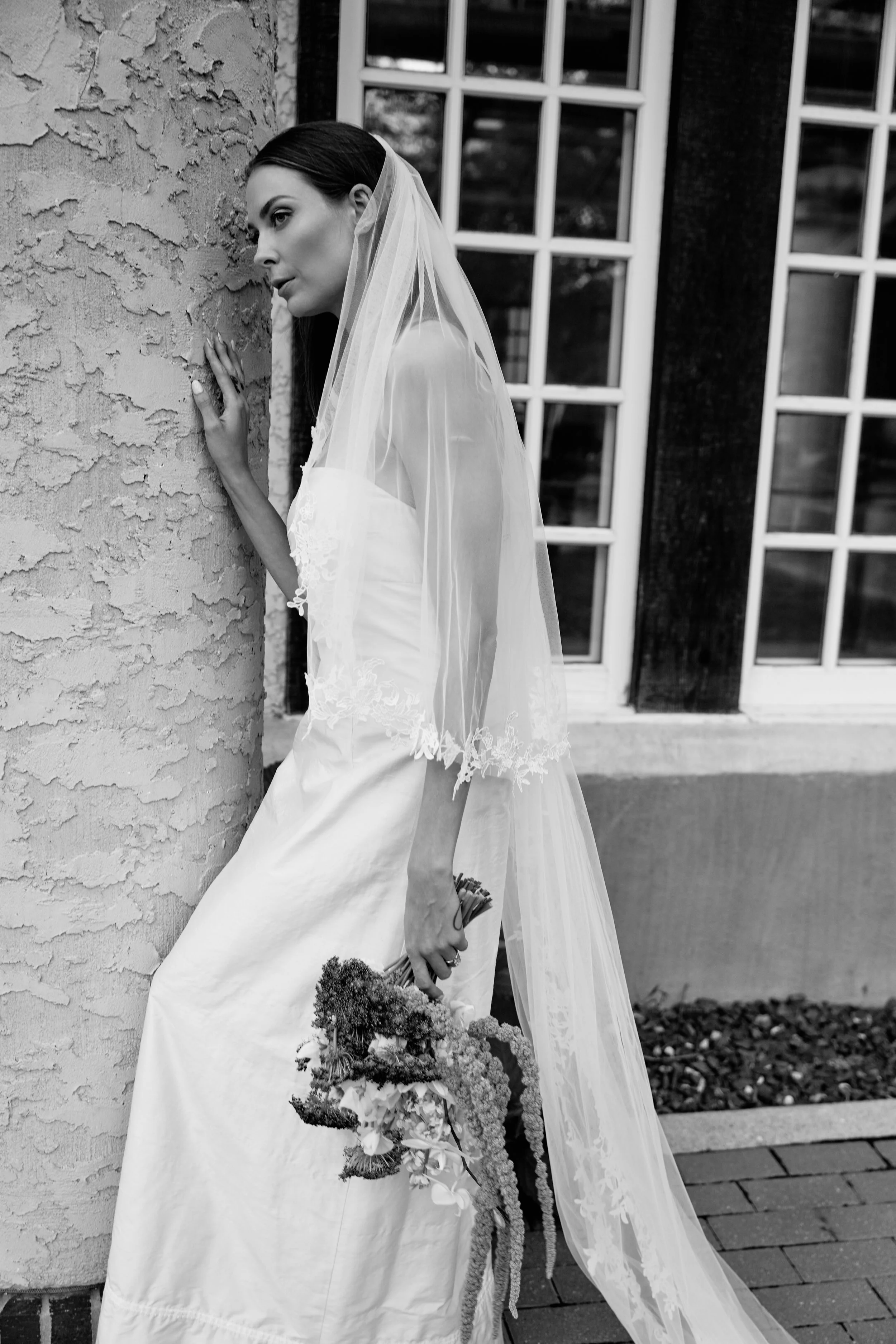Black and white photo of a woman in a wedding dress with a veil, holding a bouquet, standing outside next to a textured wall and large window.