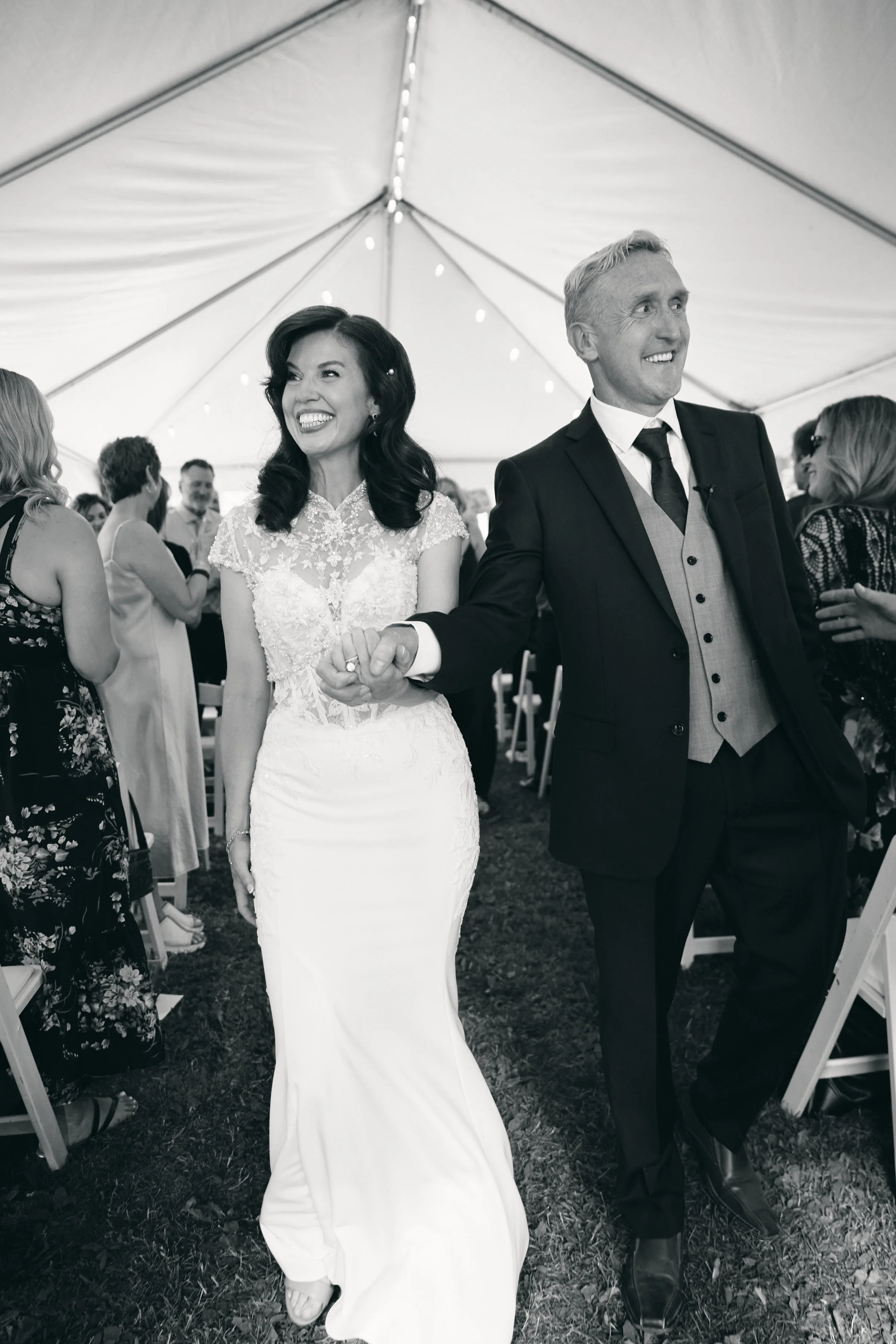 A smiling bride and groom walking hand in hand at their wedding reception under a large tent, surrounded by guests.