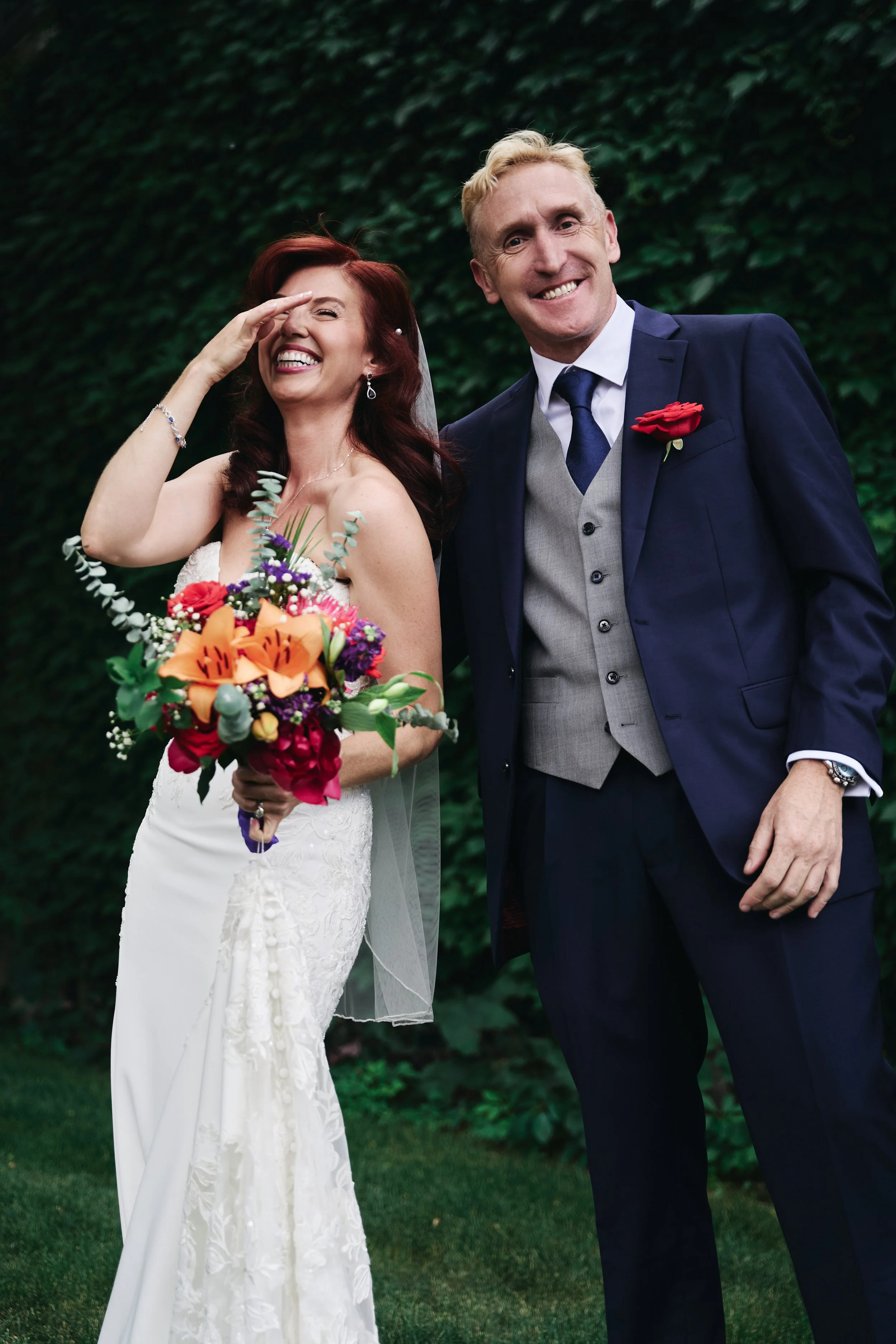 A bride and groom on their wedding day outdoors, the bride holding a colorful bouquet and the groom dressed in a navy suit with a red rose boutonniere, both smiling and laughing.