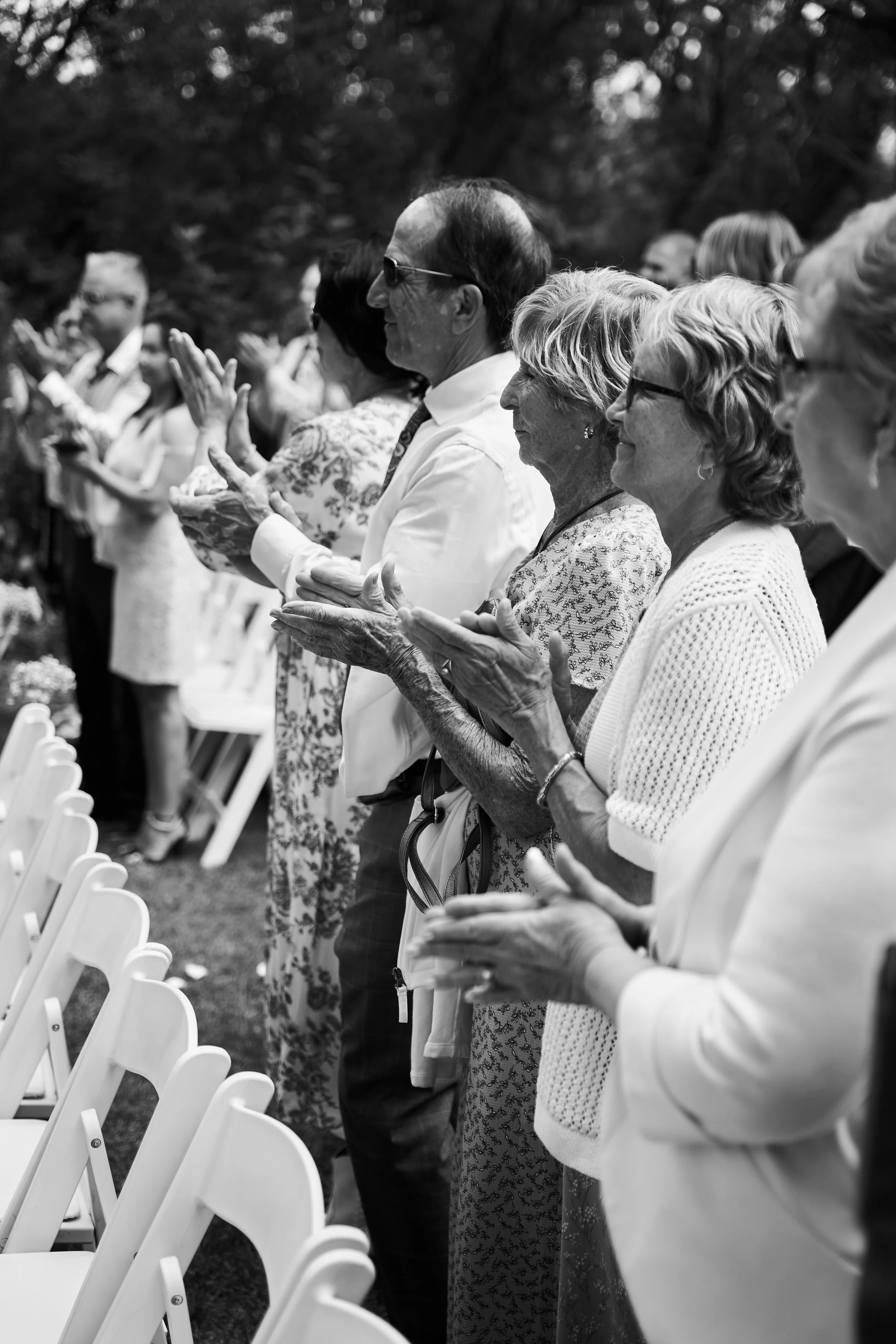 A black and white photo of a group of elderly people standing outdoors, clapping, with some seated in white chairs in front of them, at what appears to be a formal event or ceremony.