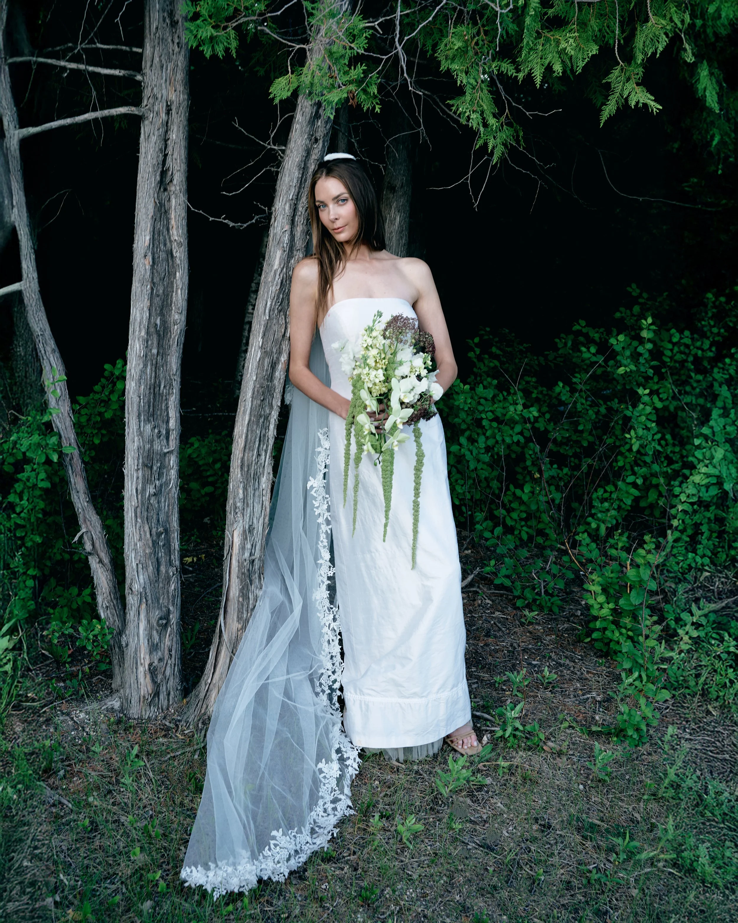 A woman in a white strapless dress holding a bouquet of flowers, standing outdoors among trees and greenery.