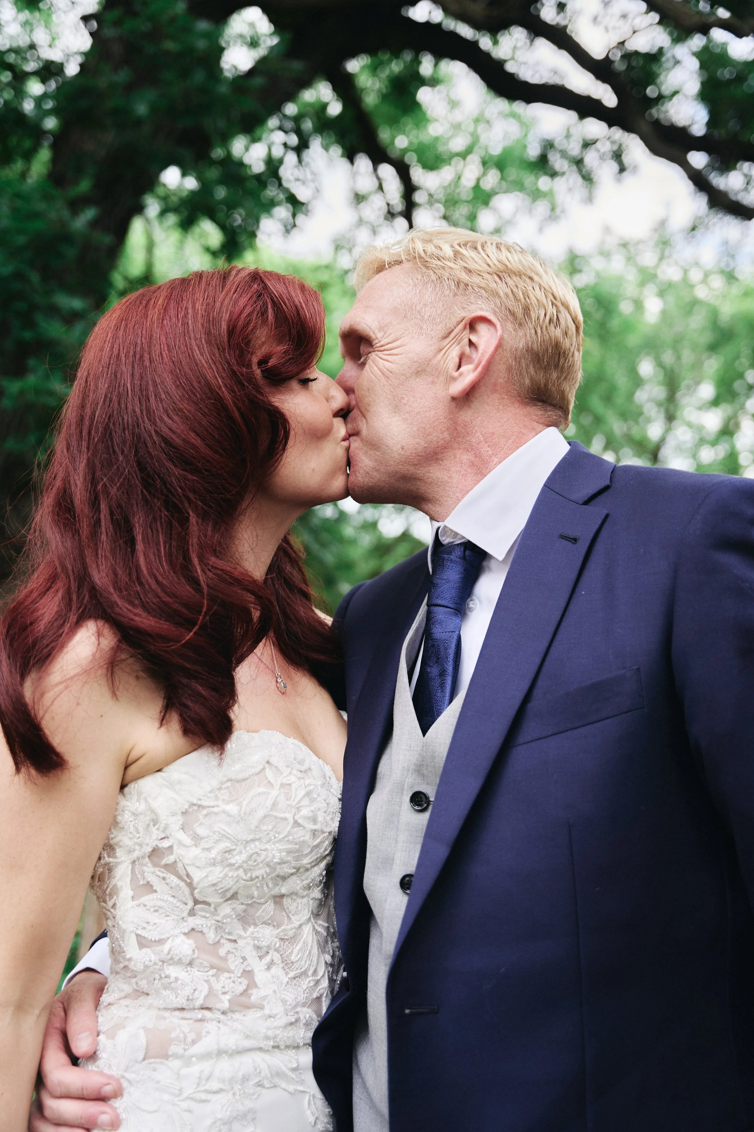 A man and woman, dressed in formal attire, sharing a kiss outdoors with trees and greenery in the background.
