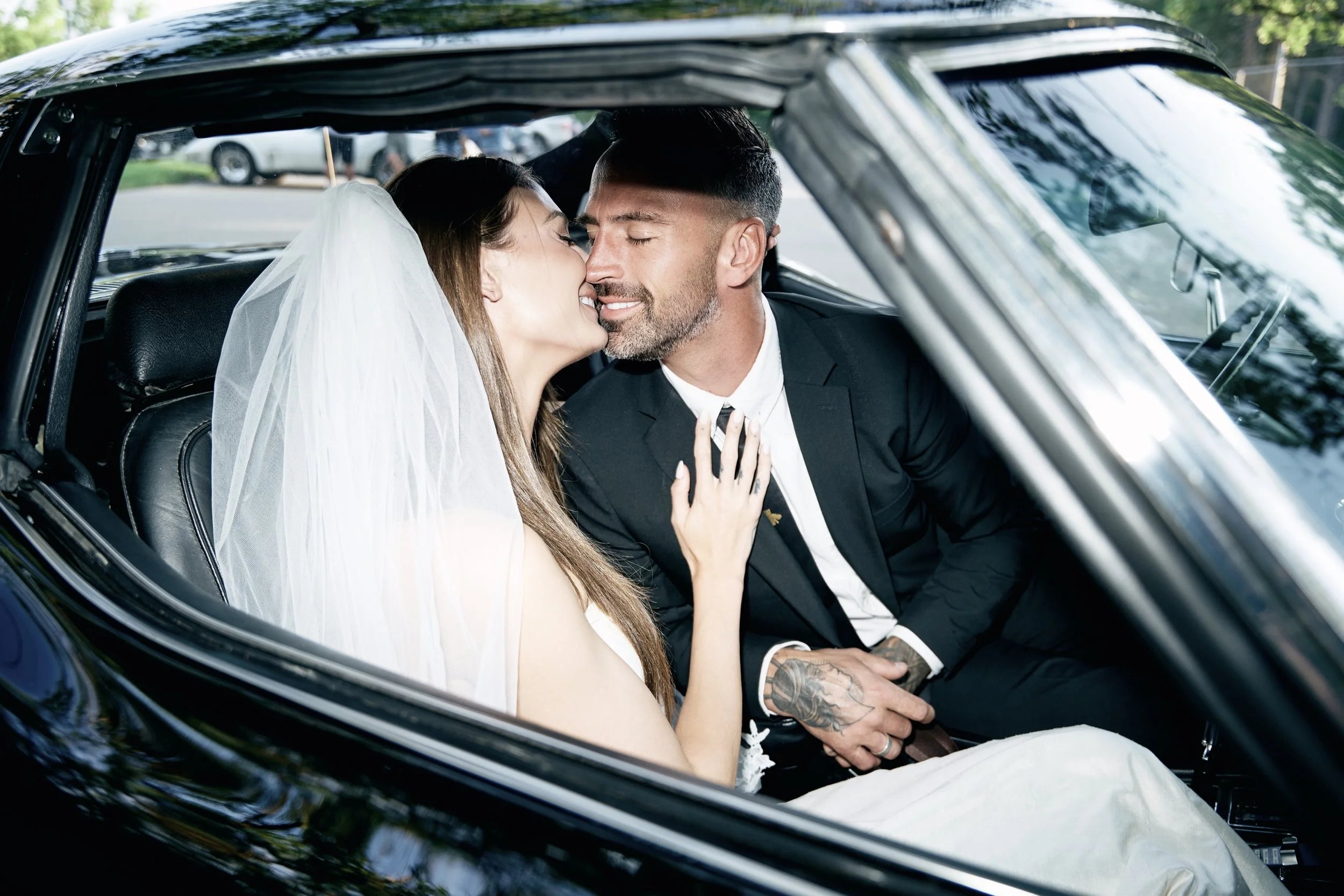 A newlywed couple sharing a kiss inside a car, with the bride wearing a veil and the groom in a black suit.