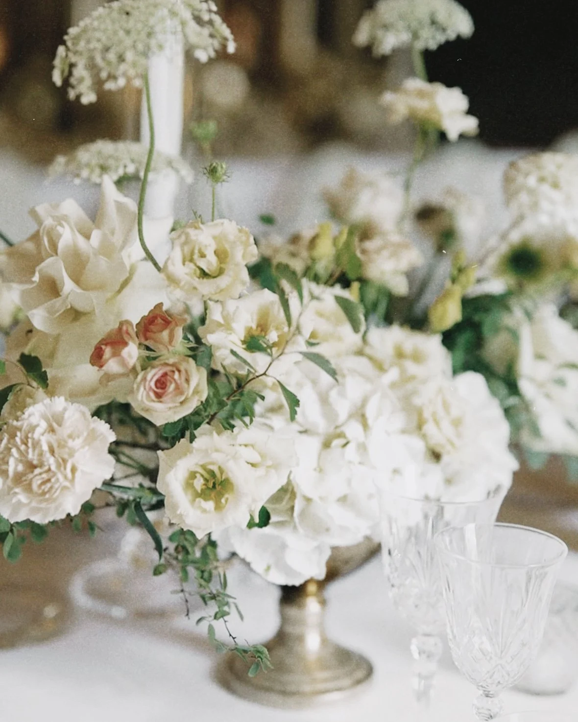 Elegant floral centerpiece with white and blush roses, hydrangeas, and greenery on a table with crystal glassware.