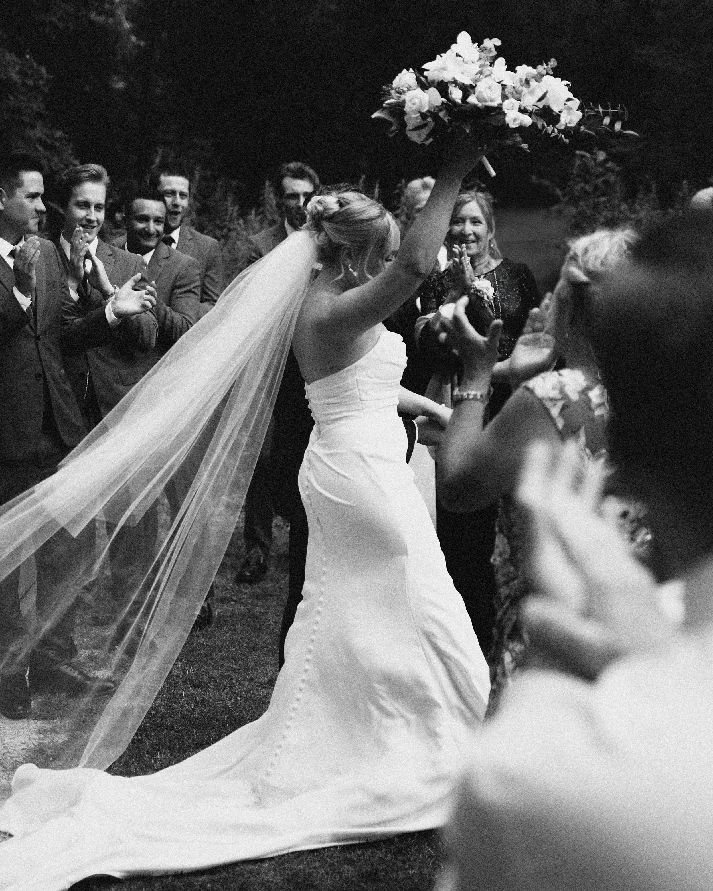 Black and white image of a bride celebrating with a bouquet of flowers at her wedding, surrounded by friends and family who are clapping and smiling.