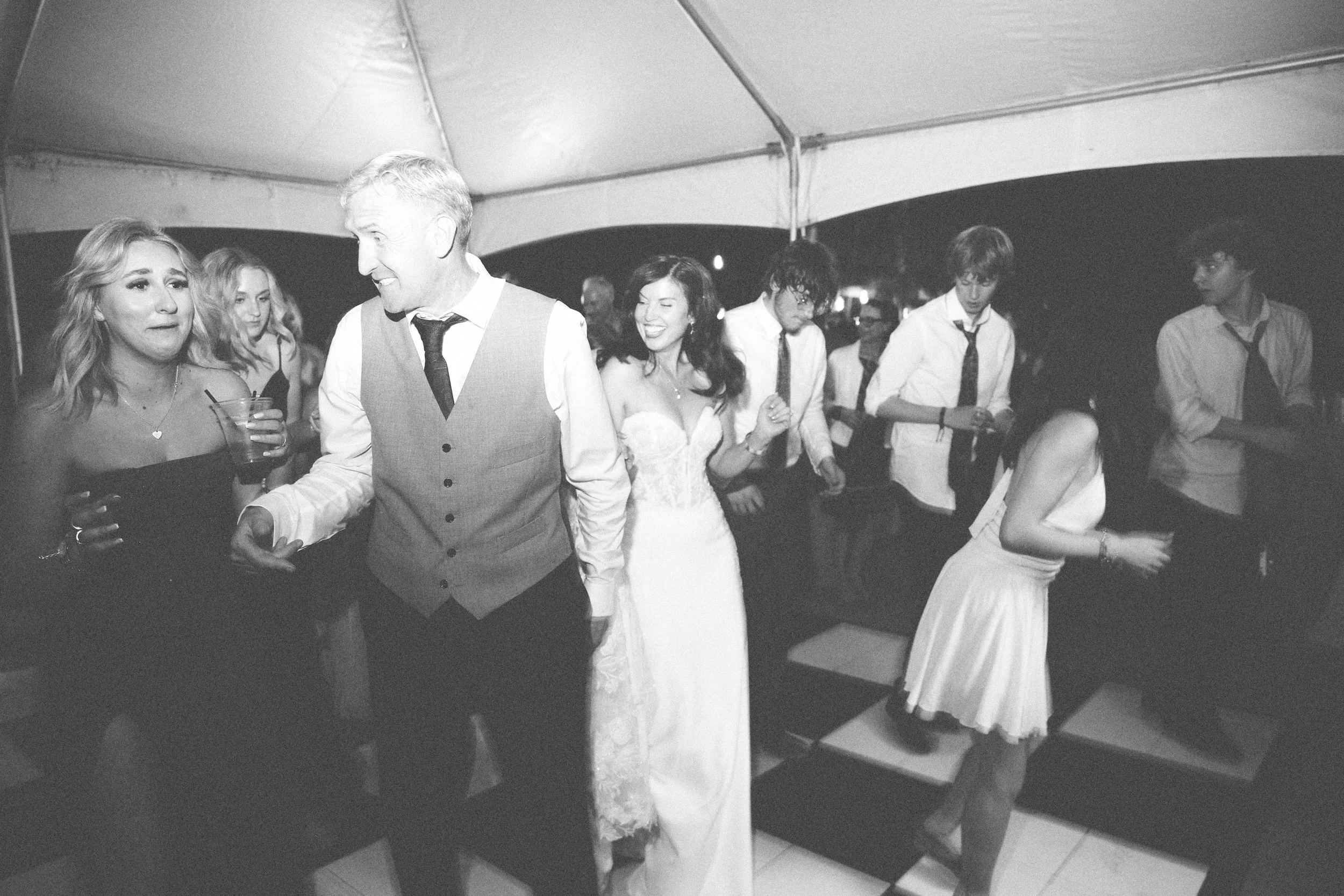 People dancing at a wedding reception inside a large tent, with the bride in a strapless wedding gown and bridesmaids in dark dresses, and groomsmen in white shirts and ties.
