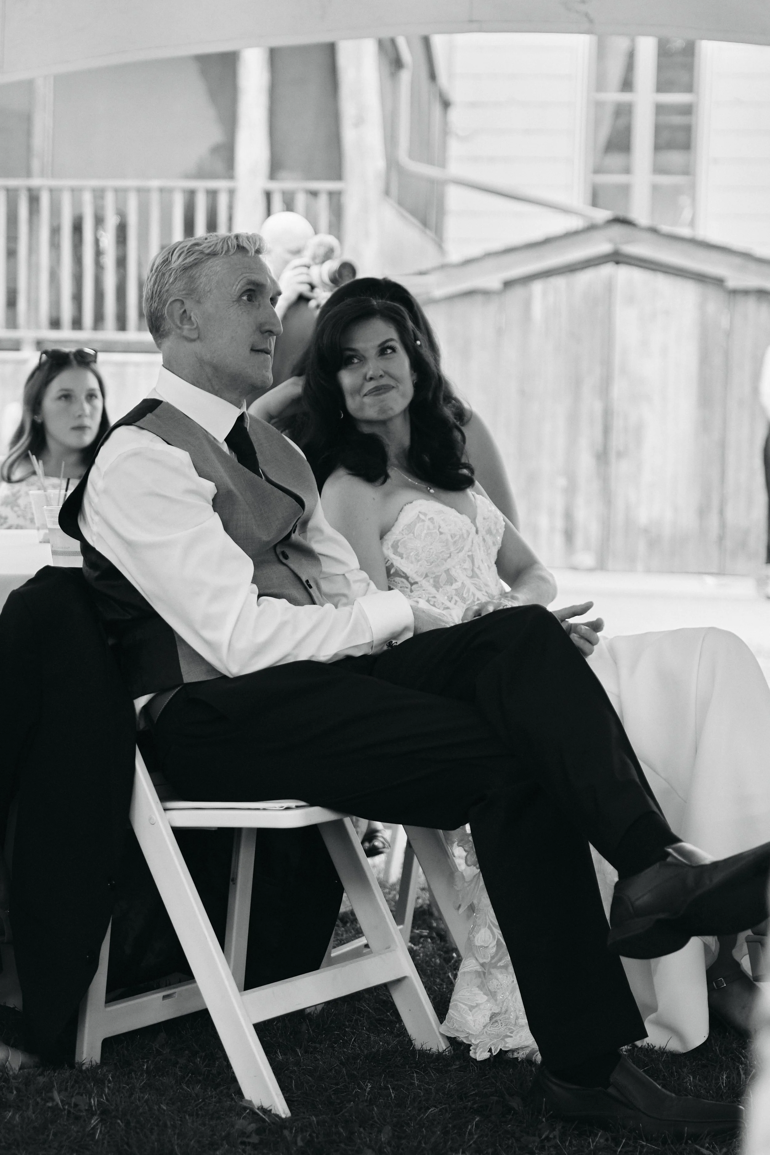 Black and white photo of a man and woman sitting next to each other at a wedding. The man is wearing a light dress shirt, a vest, and dark pants; the woman is in a white lace wedding dress. They are seated outdoors on folding chairs, with a wooden fe
