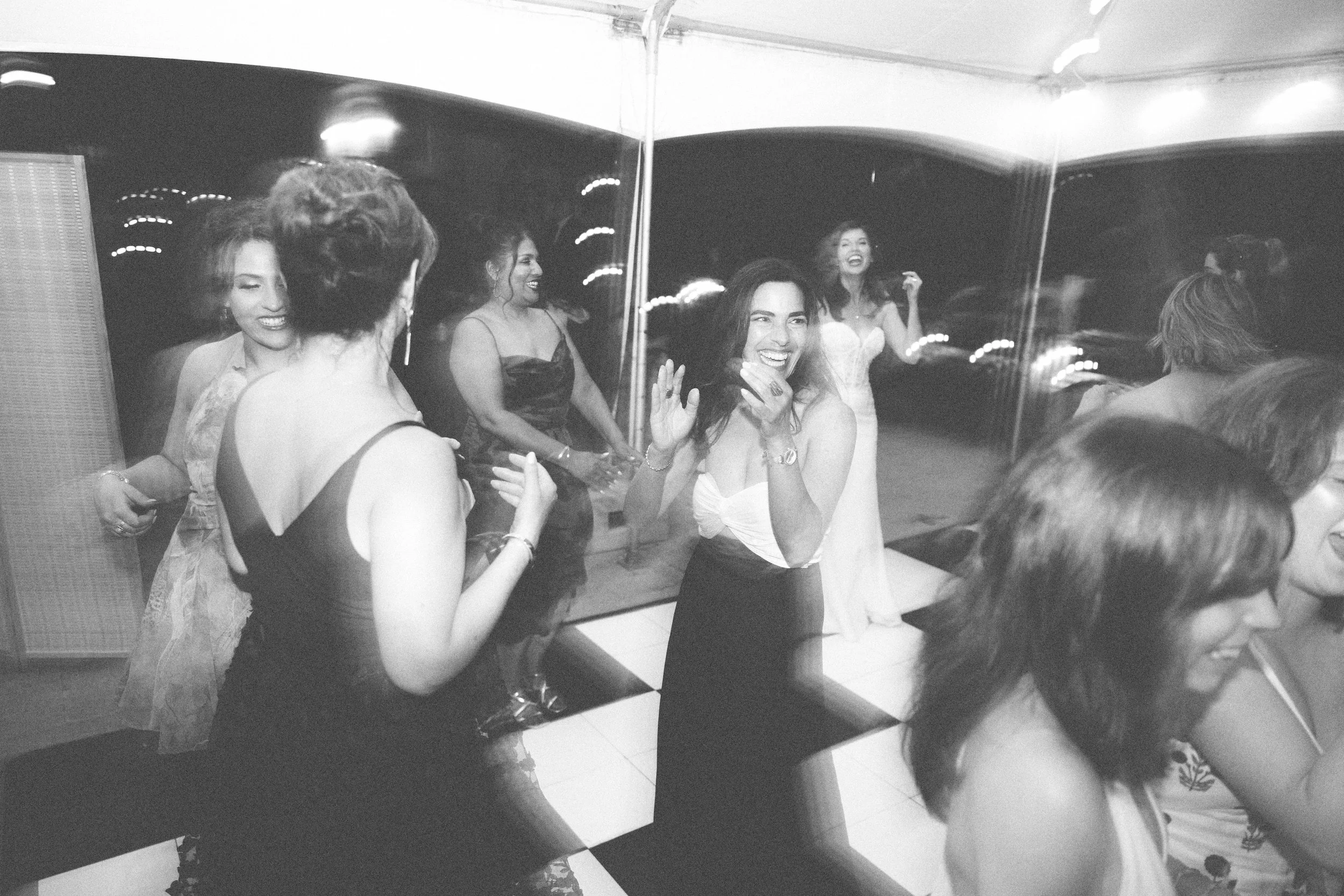Women in evening wear laughing and talking at a party with a checkered dance floor and mirrored wall.