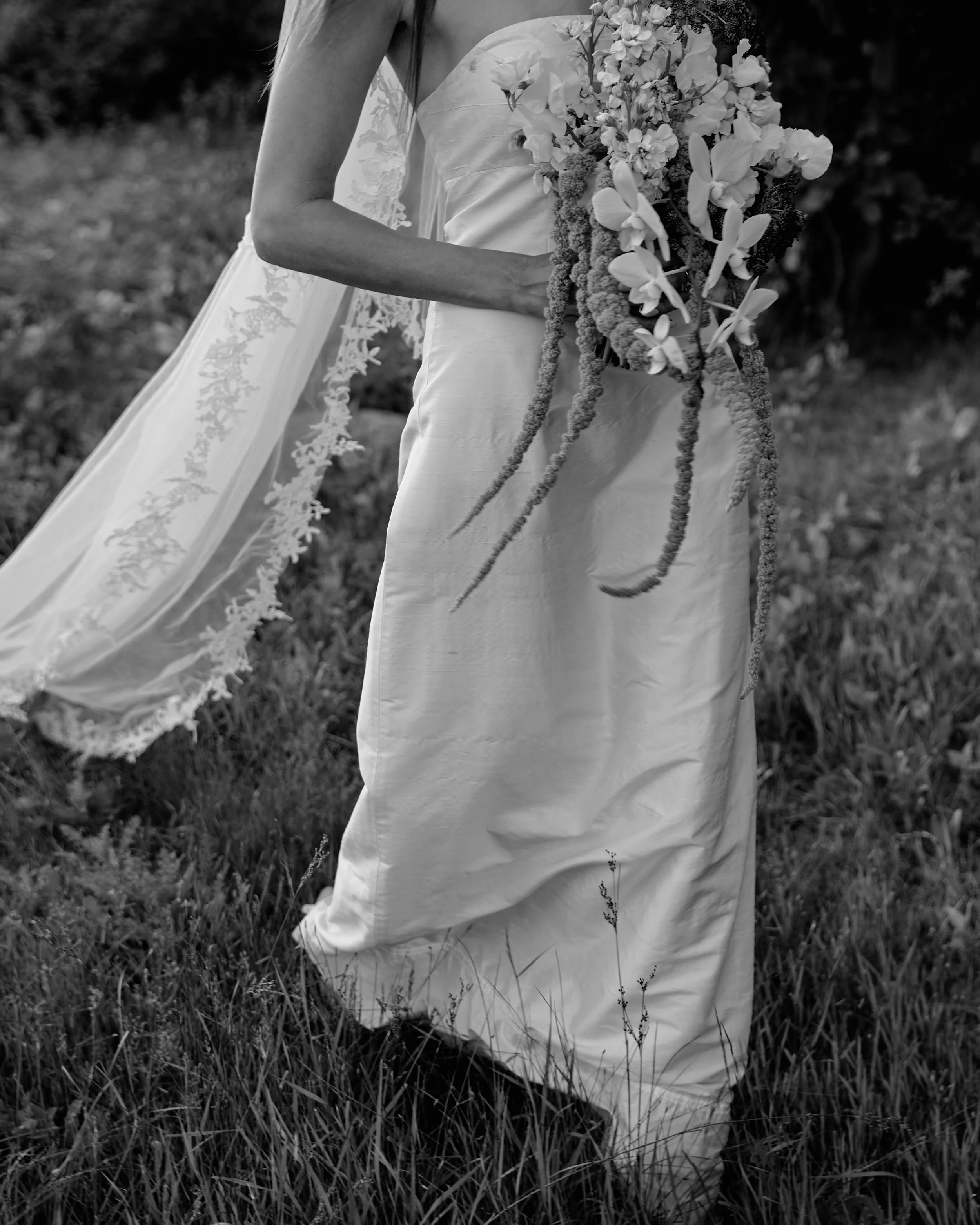 A woman in a wedding dress holding a large bouquet of flowers outdoors, with grass on the ground and trees in the background, in black and white.