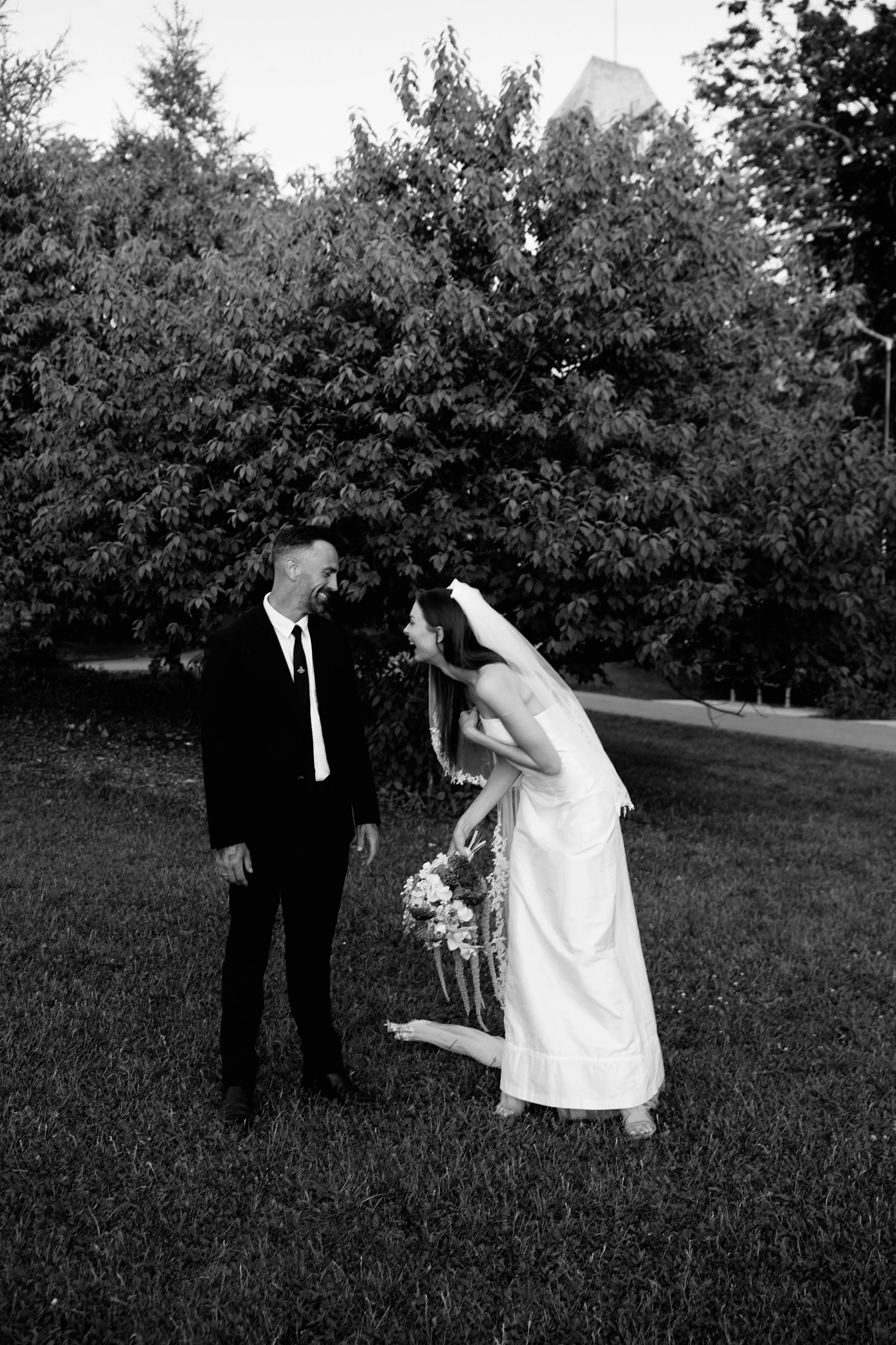 A black and white photo of a bride and a man, possibly her father, outdoors near a large tree. The bride is dressed in a white wedding gown with a veil and is holding a bouquet of flowers. She is leaning forward and laughing while the man is smiling