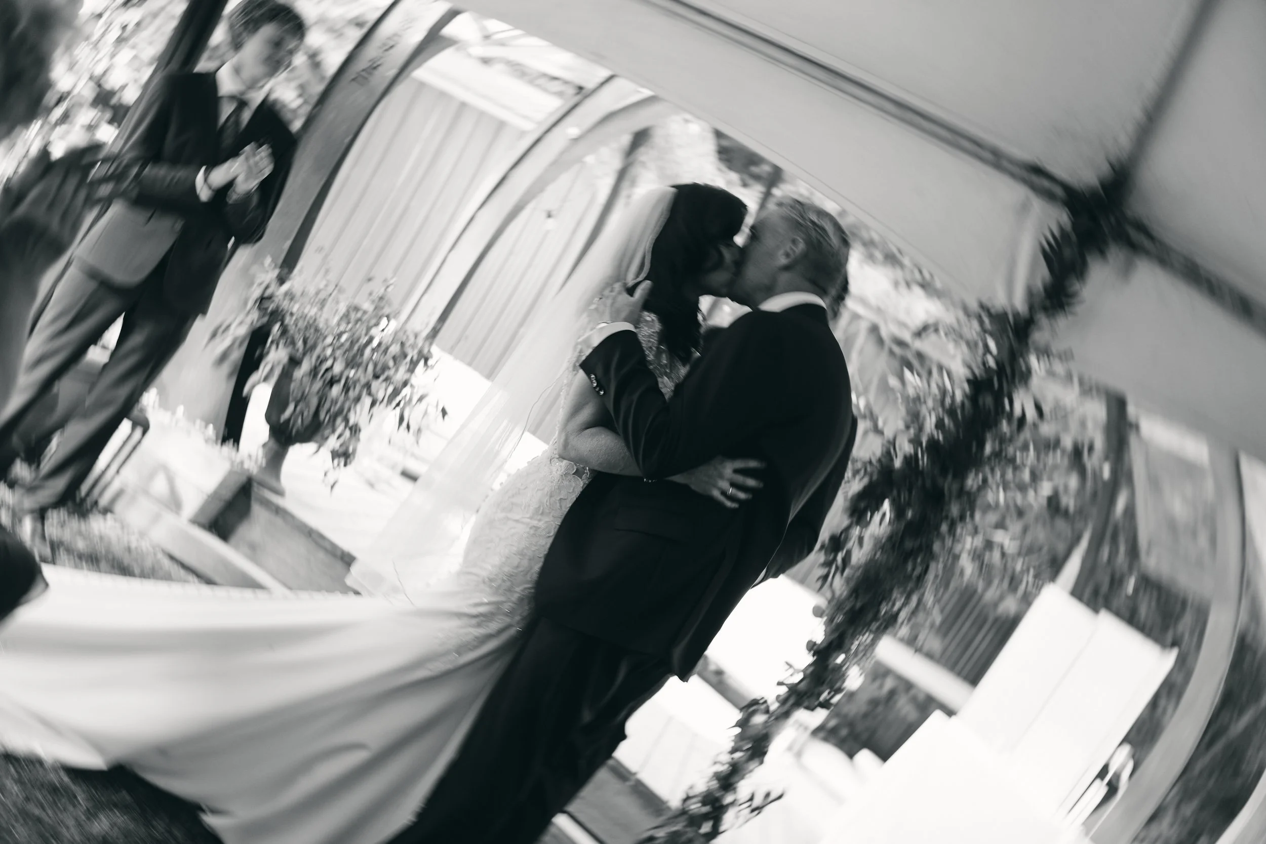 A bride and groom sharing a kiss during their wedding ceremony under a floral arch, with an officiant standing nearby in an outdoor setting.