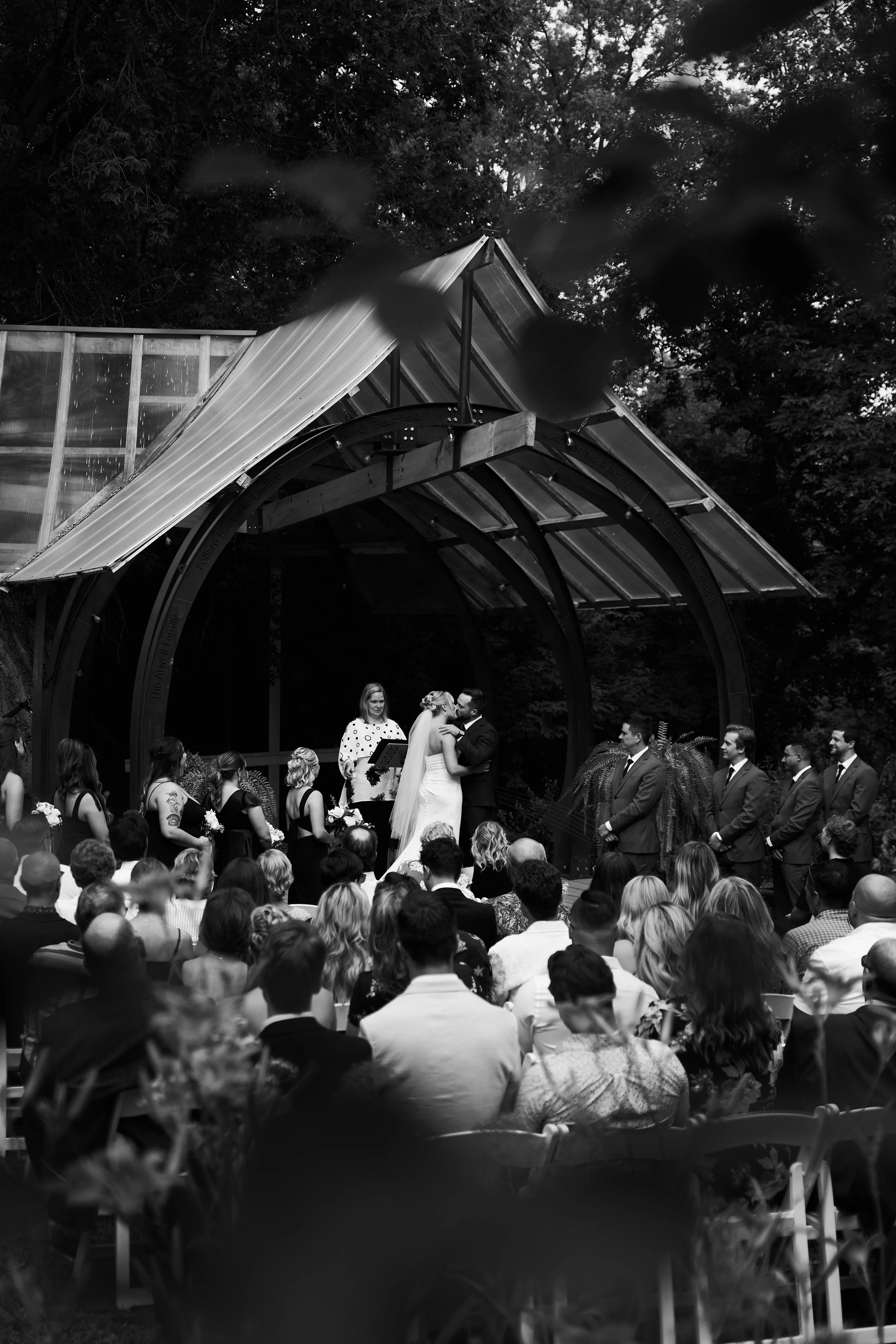 Black and white photo of a wedding ceremony outdoors with guests seated, the bride and groom kissing, officiant present, and wedding party standing nearby under a wooden and glass canopy surrounded by trees.