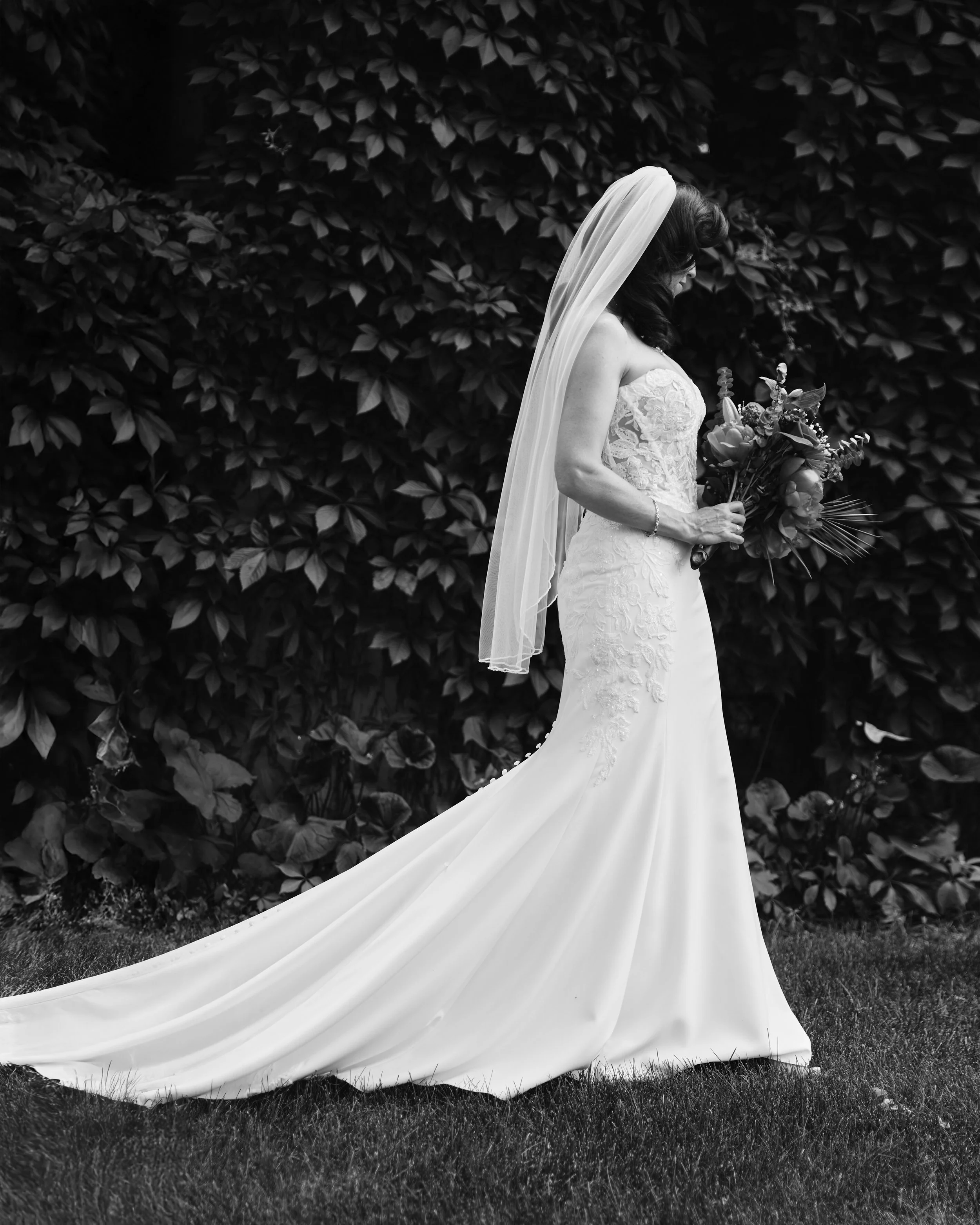 A bride standing outdoors in a wedding dress and veil, holding a bouquet of flowers, with leafy bushes in the background.