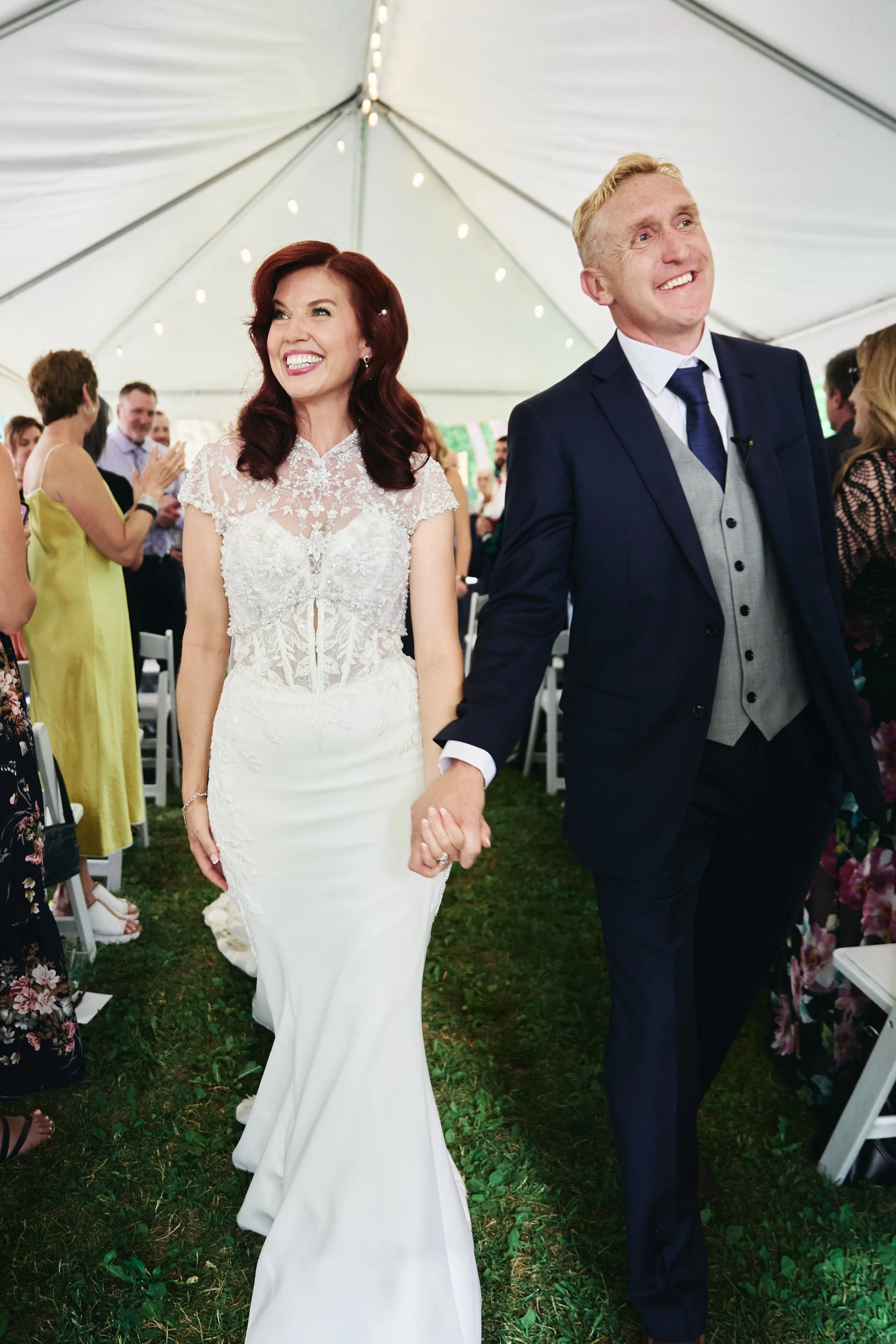 Bride and groom holding hands and smiling at a wedding ceremony under a tent, with guests standing and clapping in the background.