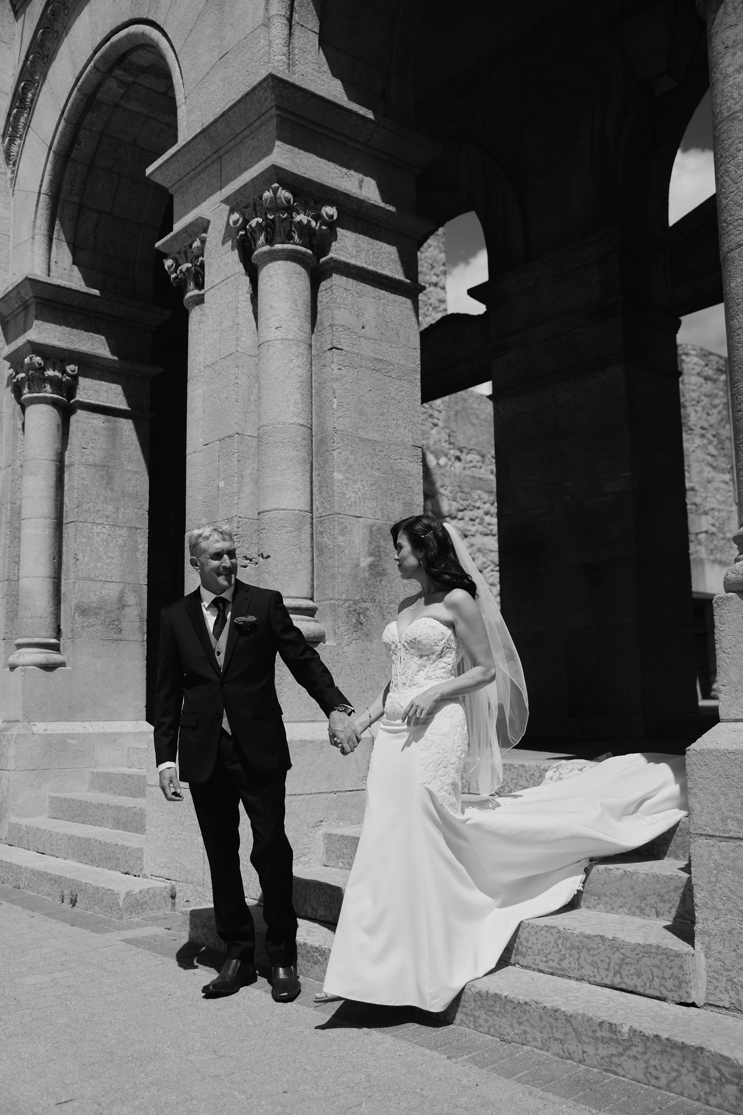 A bride and groom holding hands and smiling, standing on stone steps under an archway of a historic stone building, with the bride in a lace wedding gown and veil, and the groom in a dark suit with a tie.