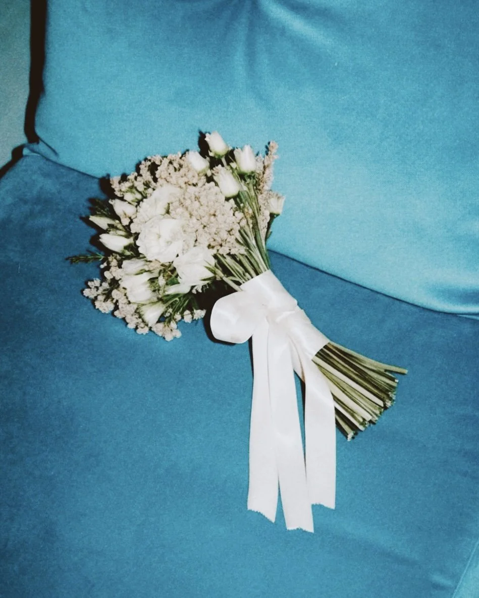 A white floral bouquet with white roses and small white flowers, tied with a white ribbon, resting on a blue fabric surface.