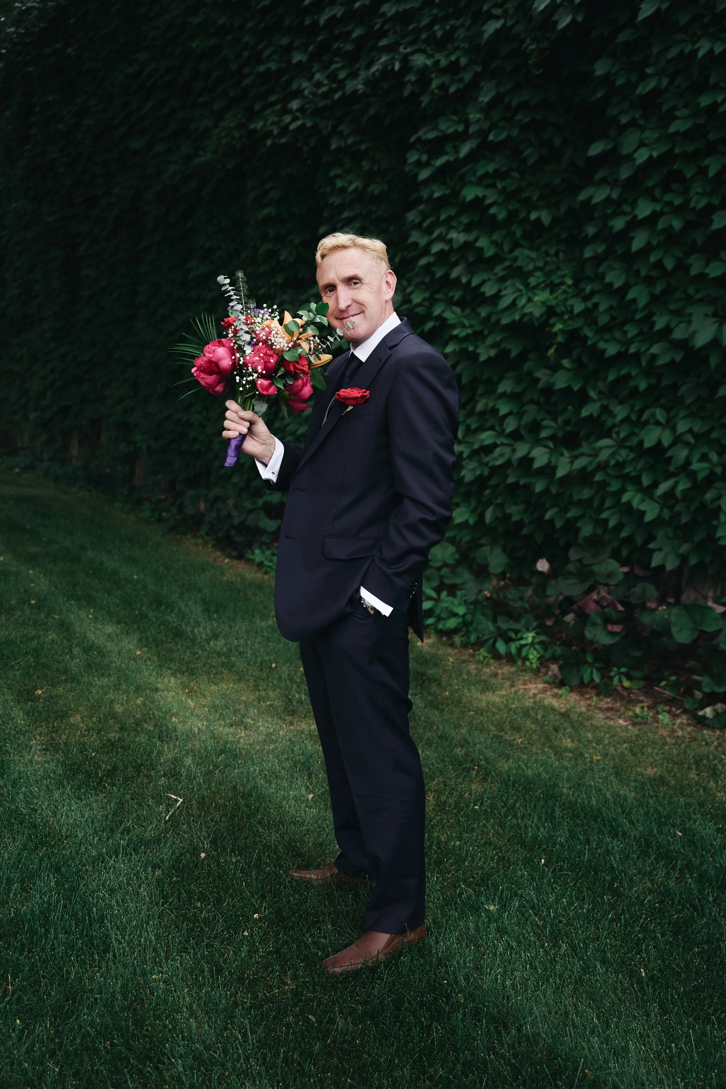 A man in a black suit holding a colorful bouquet of flowers standing on a grassy area against a background of green foliage.
