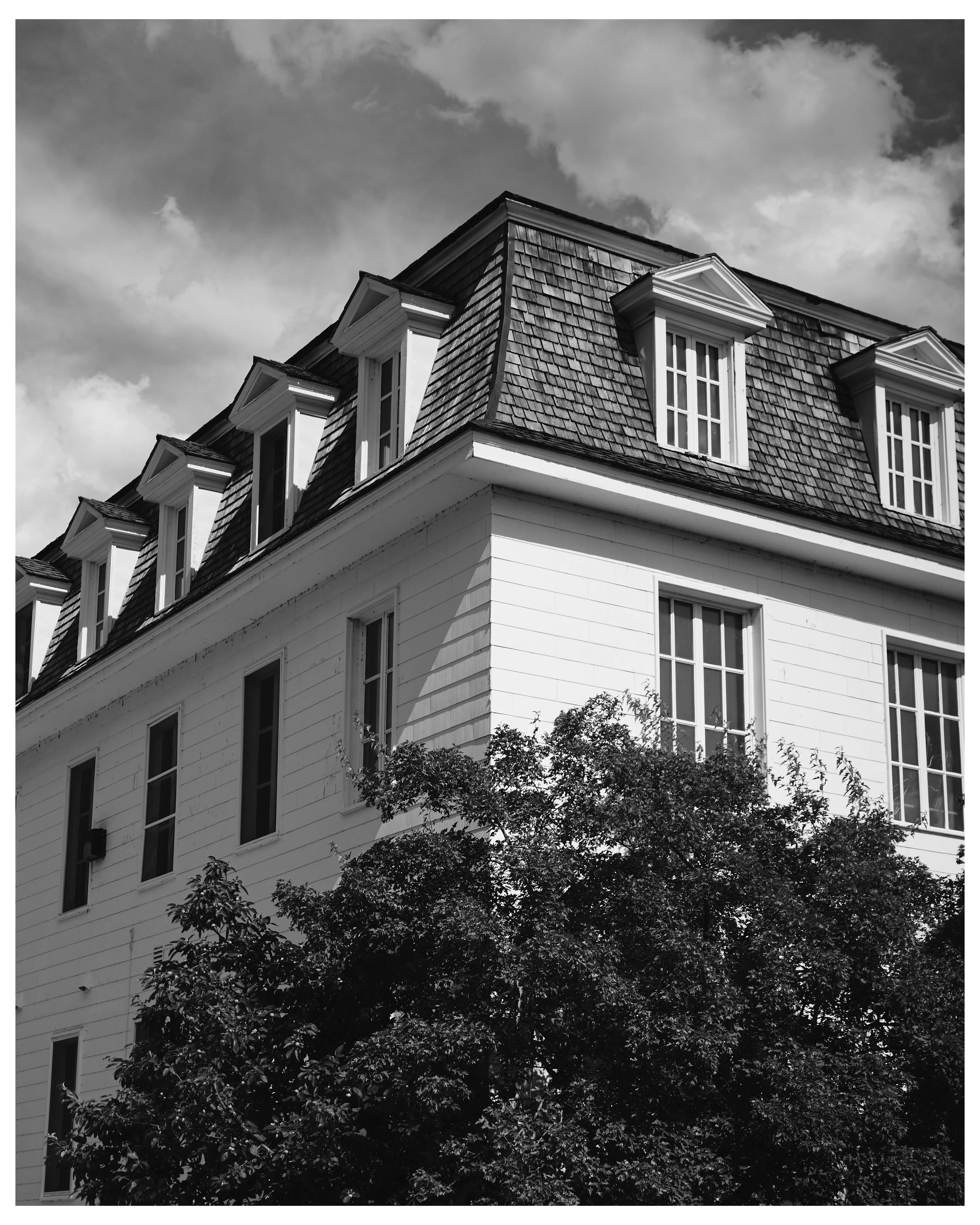 Black and white photo of a multi-story house with a steep roof, dormer windows, and a large tree in the foreground.