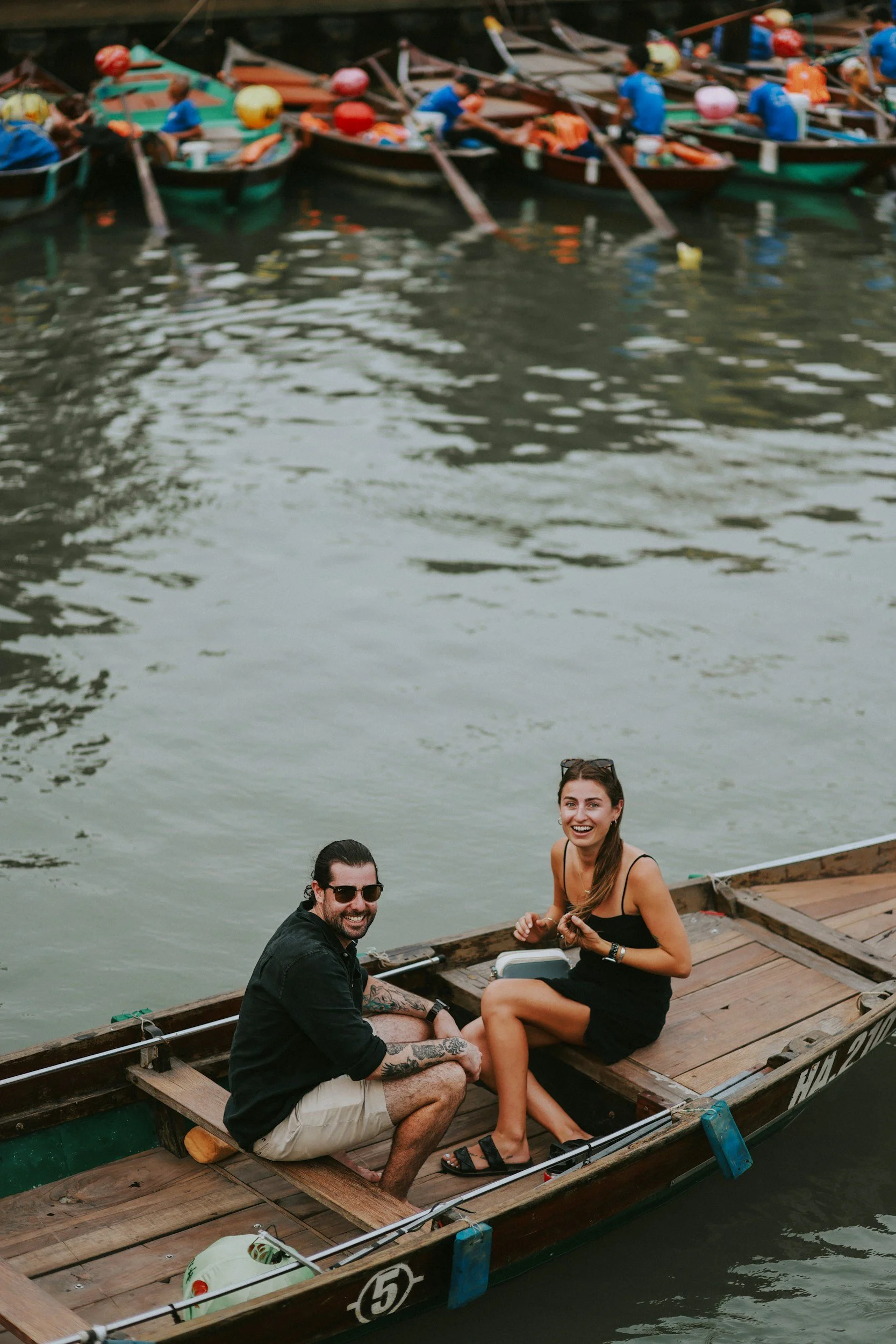 A man and a woman sitting on a wooden boat on a river, smiling and enjoying themselves, with another row of boats in the background.