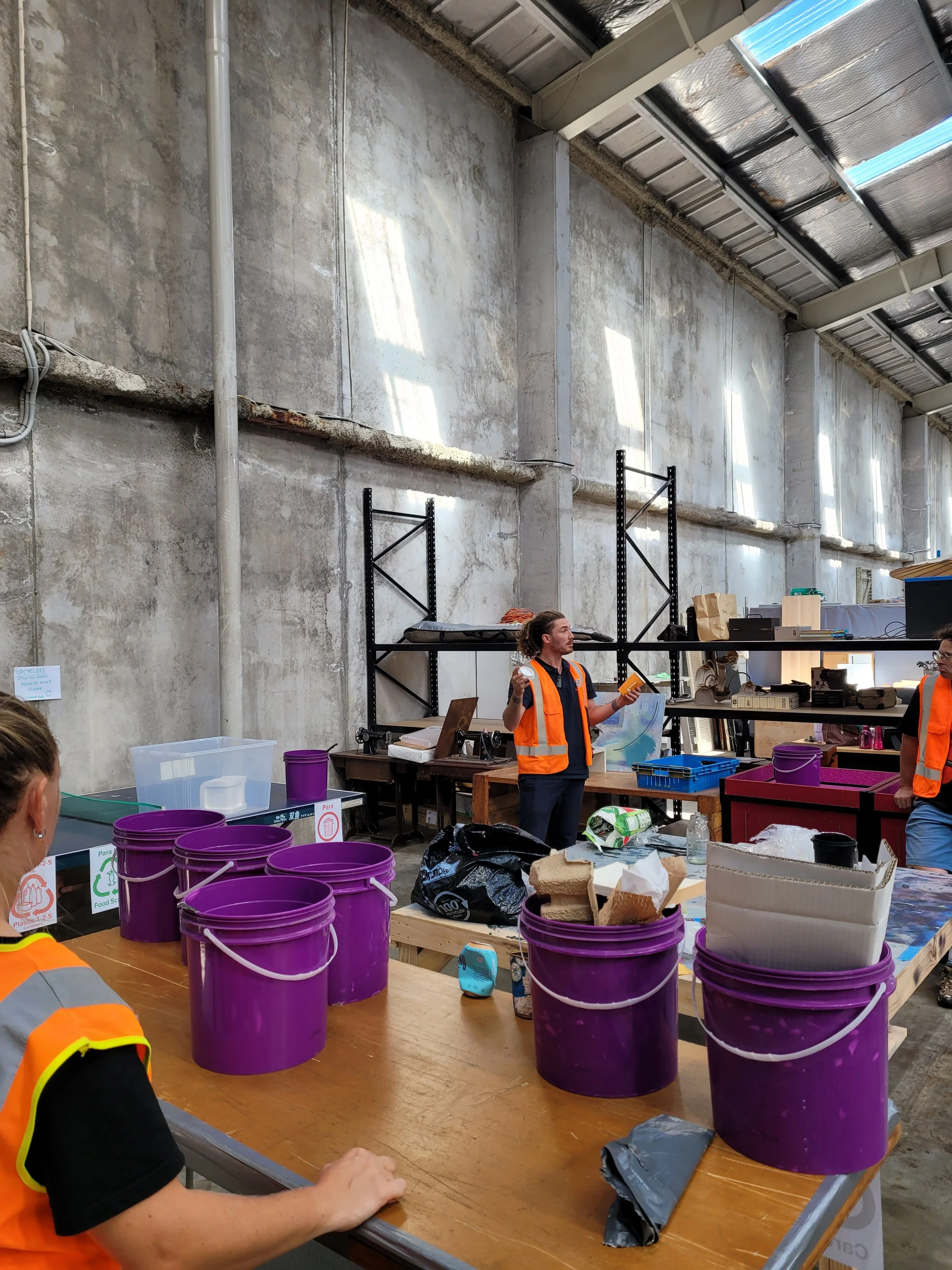 People in orange safety vests working in a warehouse or industrial space with high ceilings, concrete walls, and large purple buckets on a table.