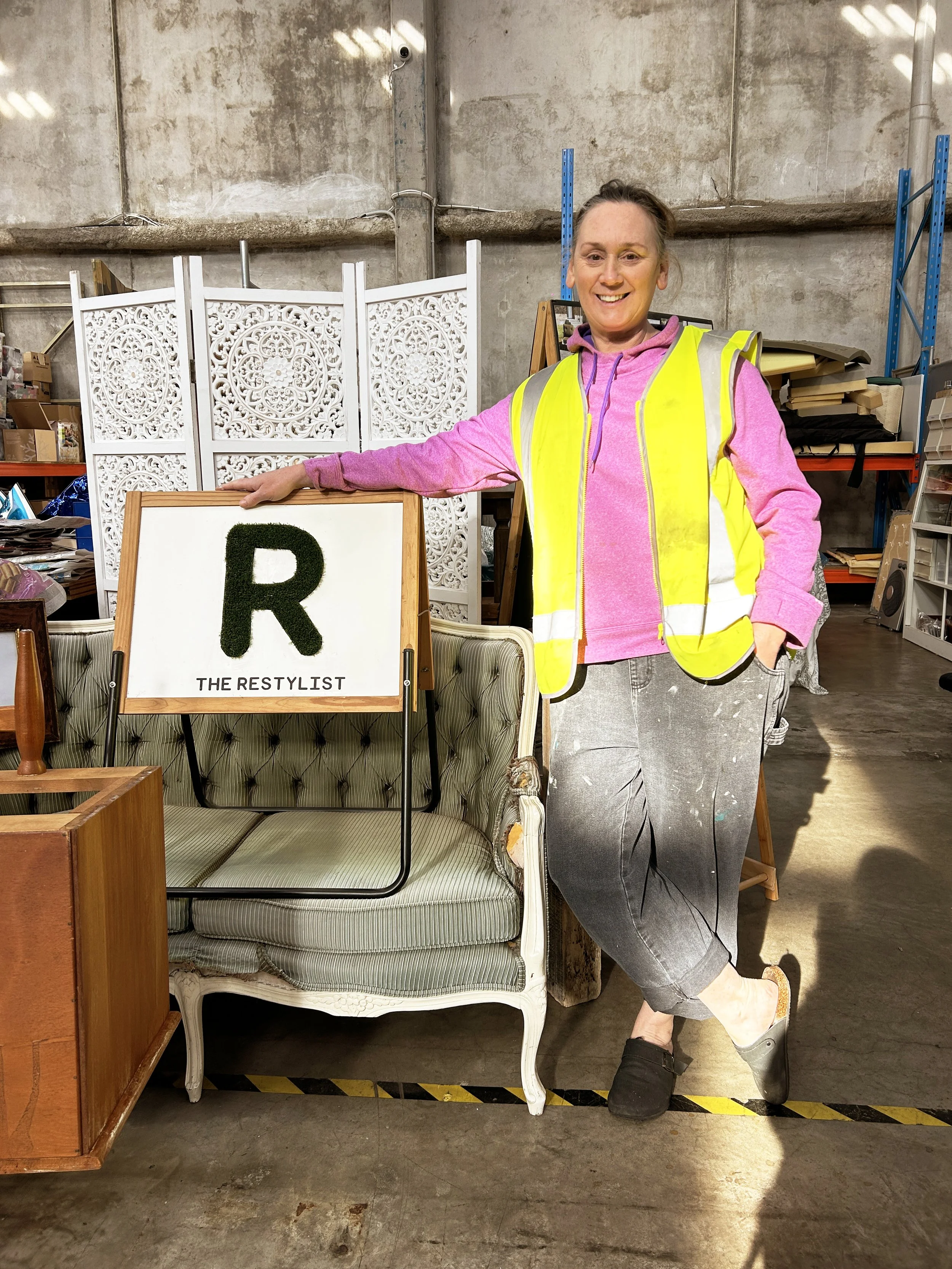A woman in a pink hoodie and black slippers wearing a yellow safety vest, smiling and standing next to a decorated sign that reads, 'R THE RESTYLIST.' She is in an industrial-style space with shelves and furniture in the background.