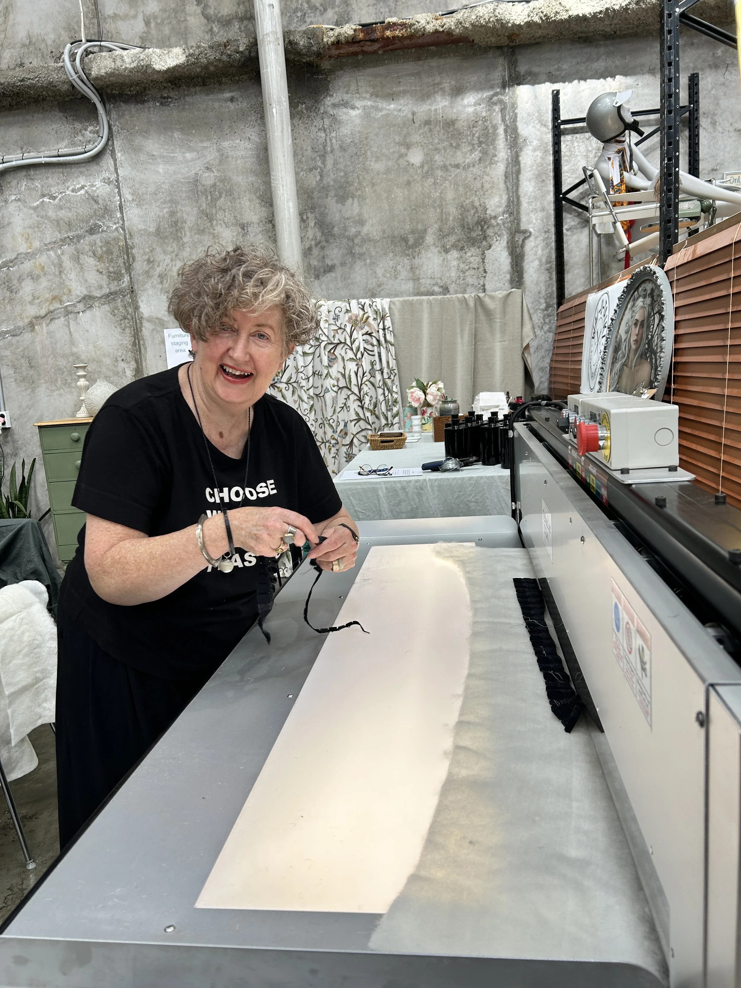 A woman with curly gray hair smiling and working on a project at a stainless steel table with a large sheet of paper, in a room with concrete walls and shelves. There is a mannequin wearing a helmet and a scarf on the shelf behind her.