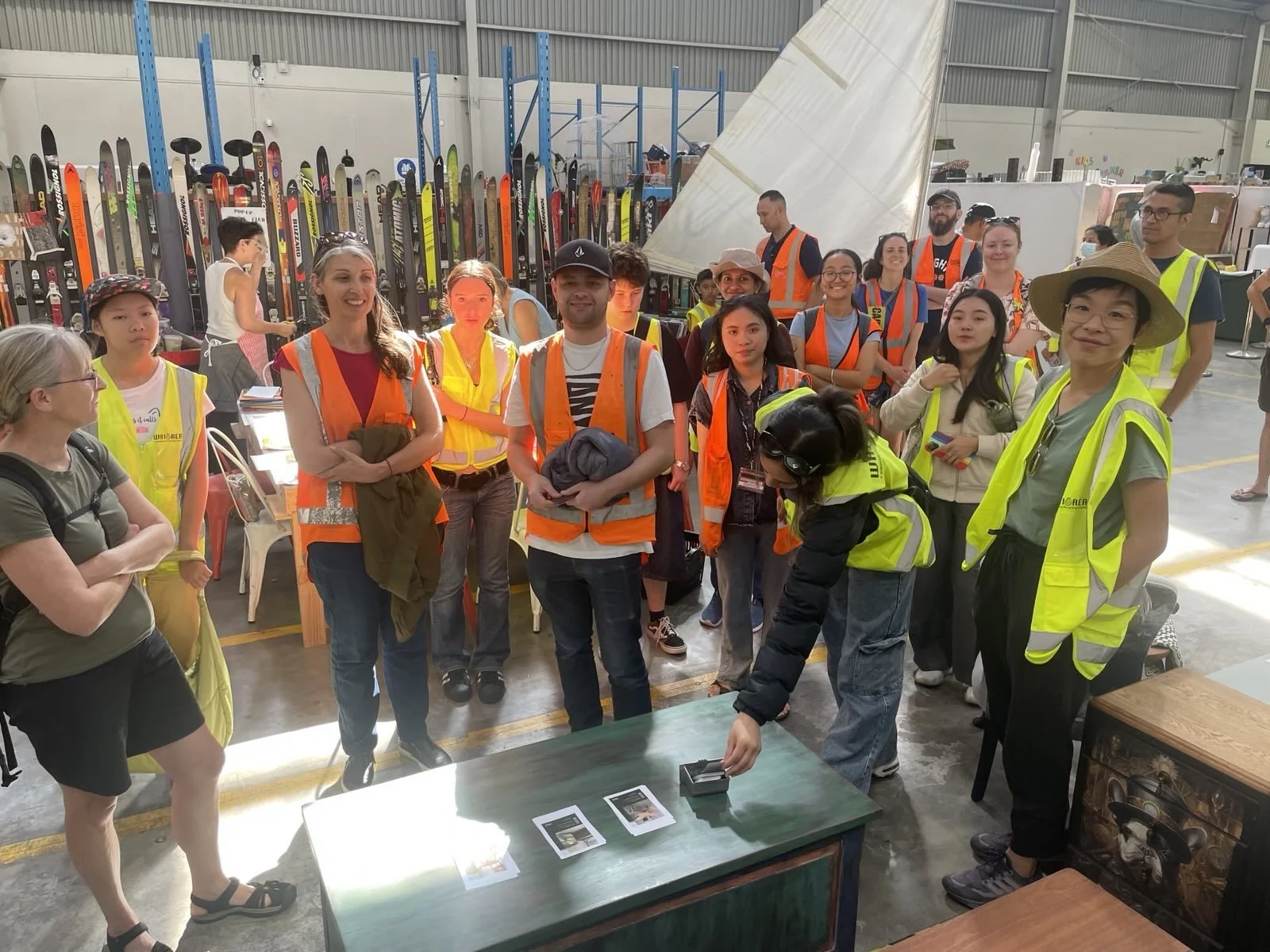 A group of people wearing safety vests gathered around a woman in safety gear who is arranging items on a table in a warehouse filled with skis and equipment.