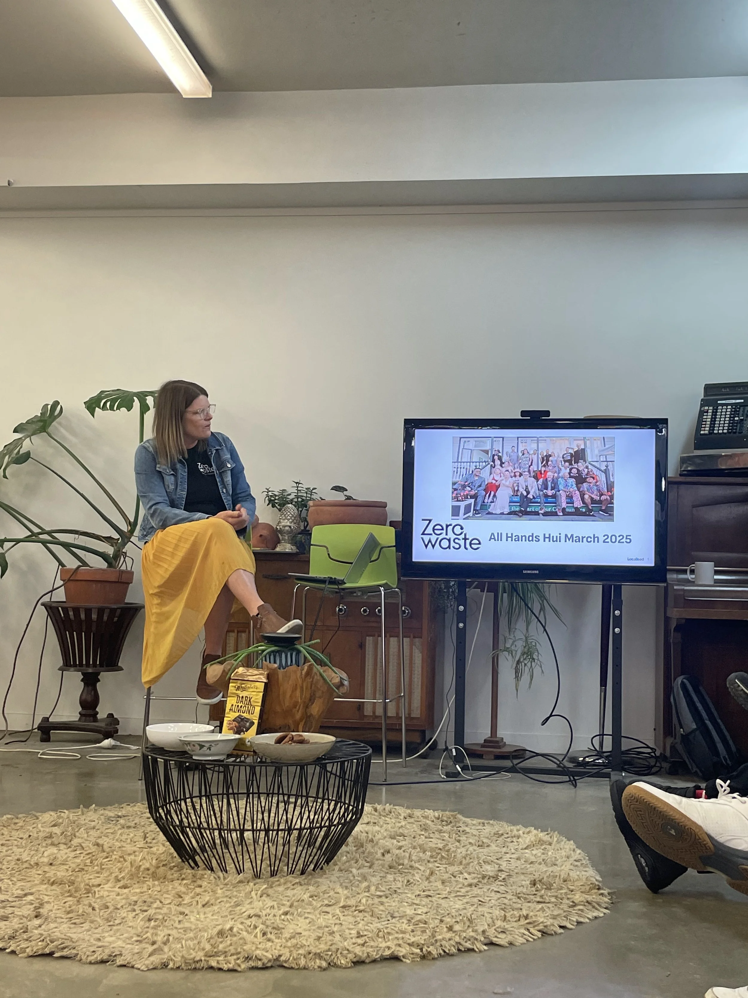 A woman sitting on a stool gives a presentation on Zero Waste at All Hands Hui March 2025, with a TV displaying the event details. The room has plants, a rug, and a coffee table with snacks.