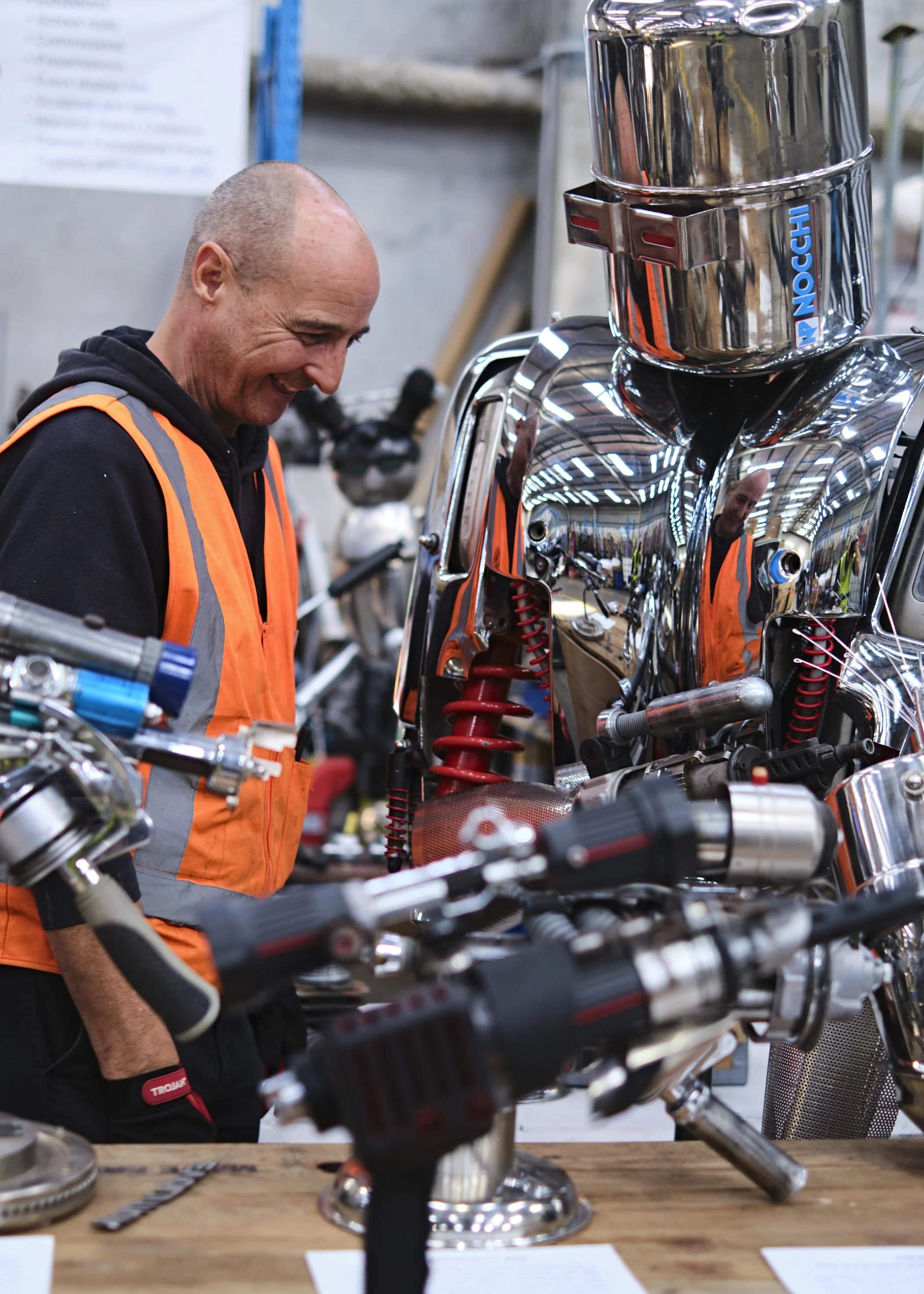 A man in an orange safety vest working on a chrome motorcycle frame in a workshop.