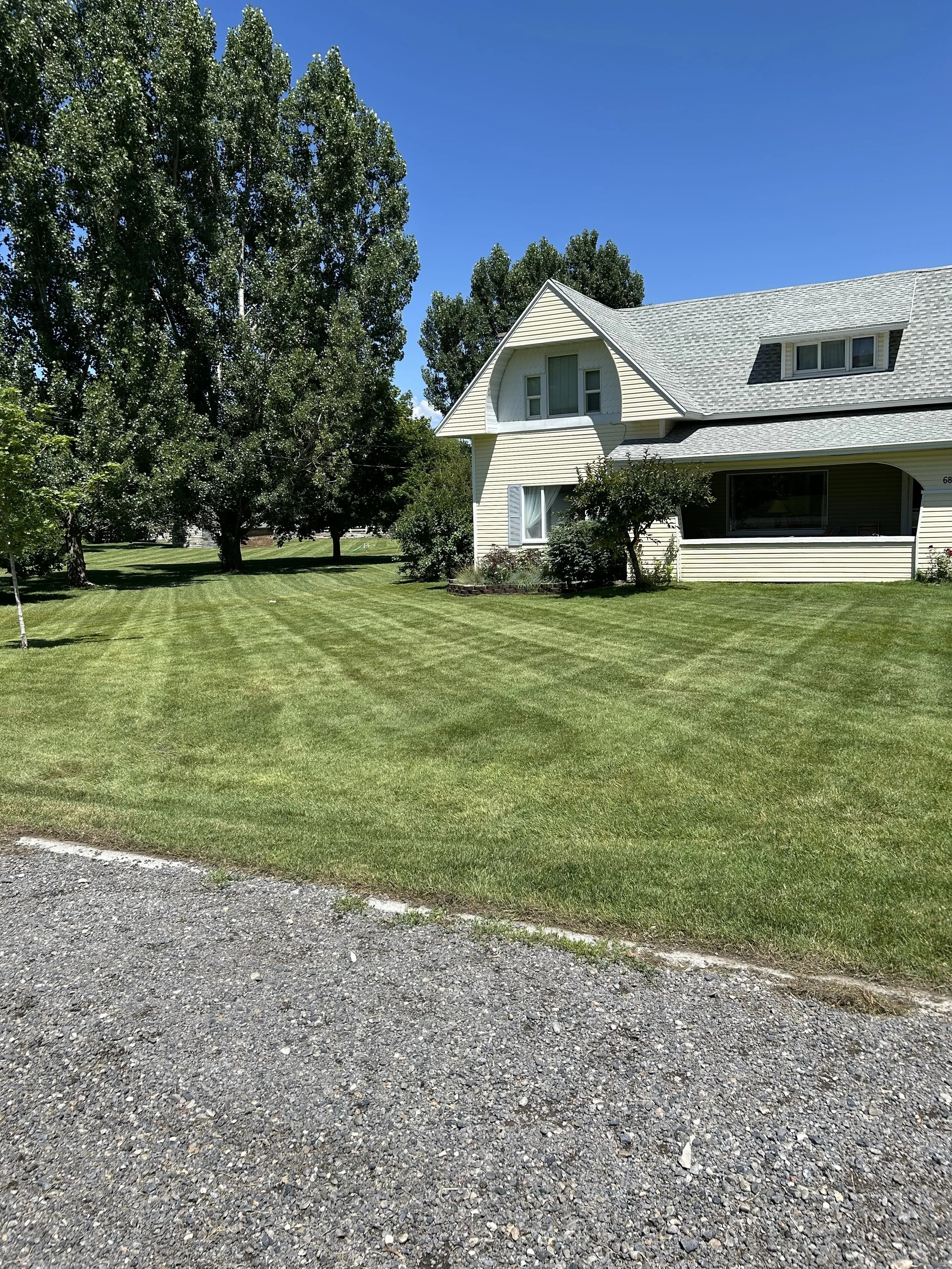 A residential house with beige siding and a gray roof, surrounded by a well-maintained green lawn and trees, under a bright blue sky.
