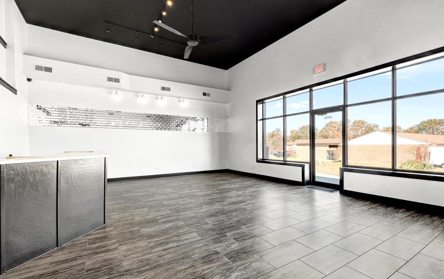 Empty commercial space with large windows, dark ceiling fan, wood and tile flooring, and a partial wall with mirror accents.