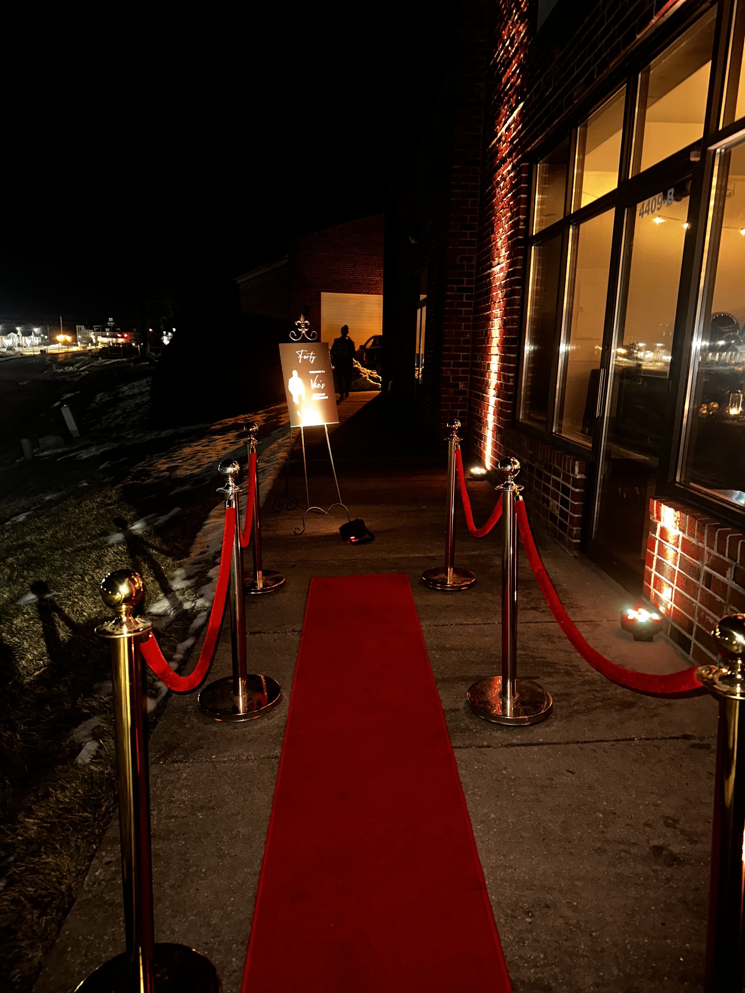 Red carpet entrance with red velvet ropes leading to a building at night, illuminated by warm lighting with a sign on a stand nearby.