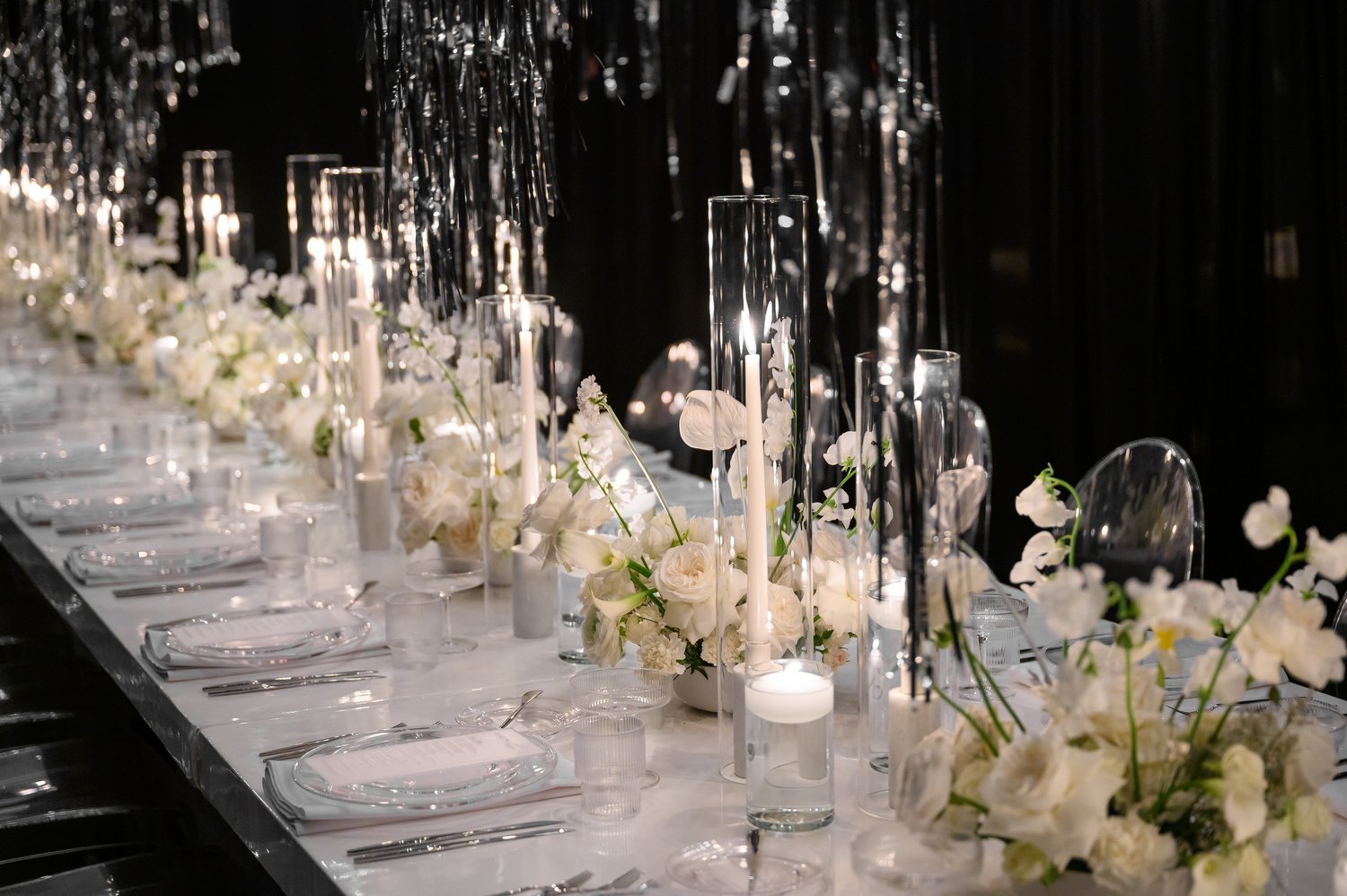 Elegant wedding reception table decorated with white flowers, tall glass candle holders with lit candles, and hanging silver streamers.