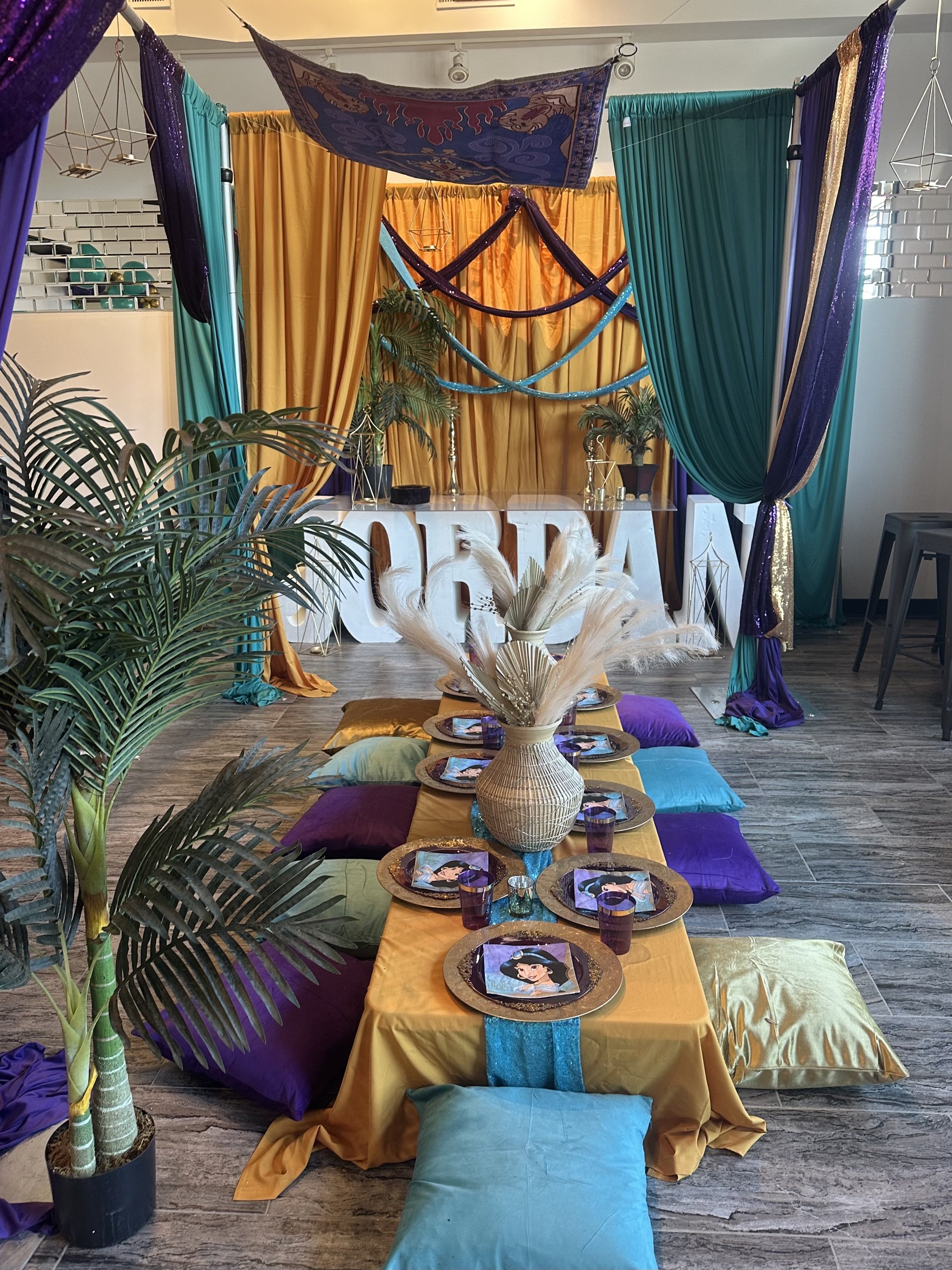 Colorful party setup with a low table covered with a gold tablecloth, decorated with a vase of pampas grass, surrounded by assorted colorful cushions. Behind, a draped backdrop with curtains in teal, purple, and gold, adorned with hanging geometric d